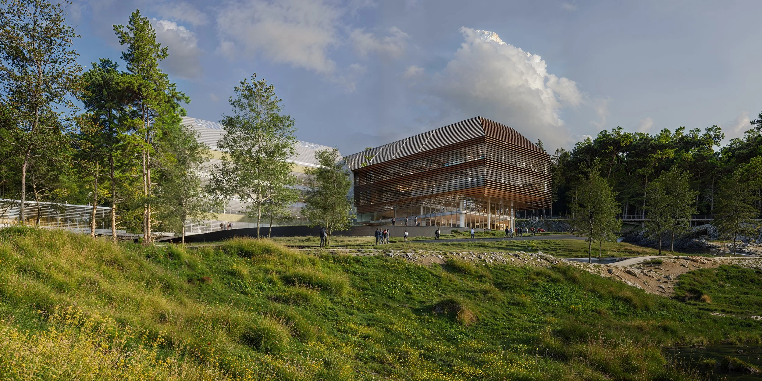 Modern building with glass walls and wooden slats, surrounded by green trees and grassy landscape, under a partly cloudy sky.