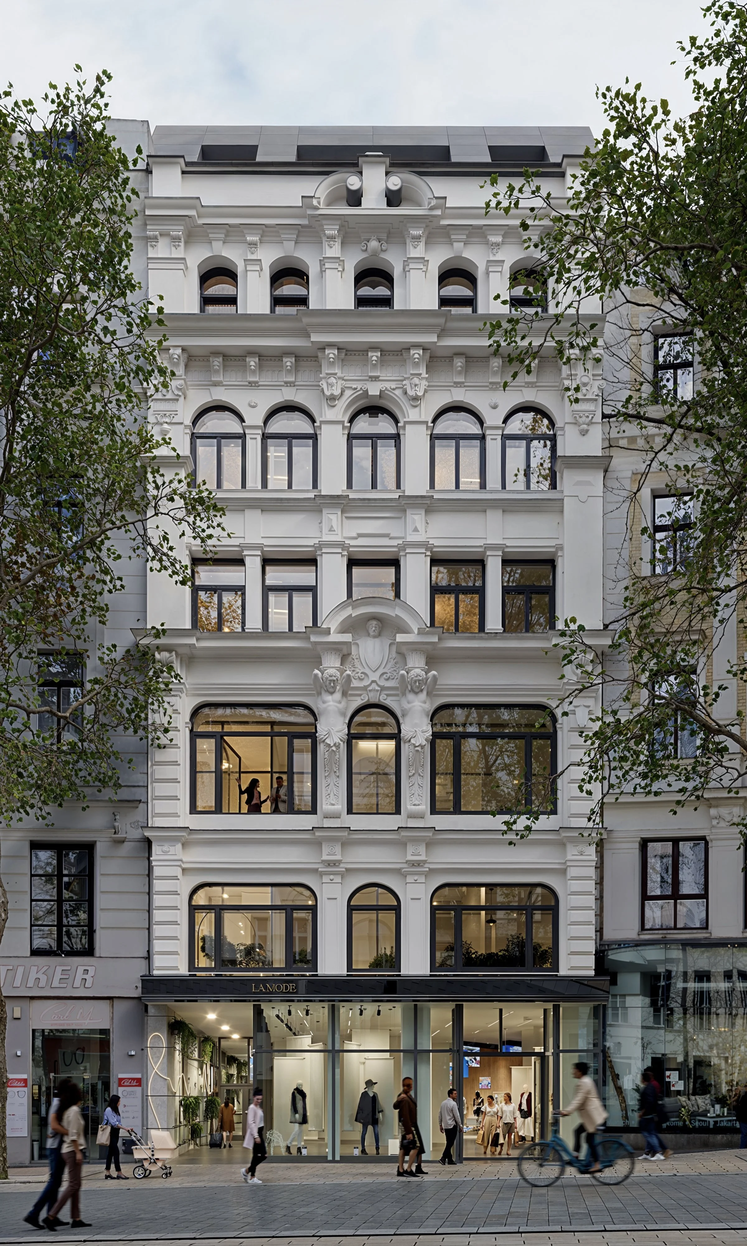 Multi-story white building with ornate architectural details, large windows, and a commercial space on the ground floor with pedestrians walking by.