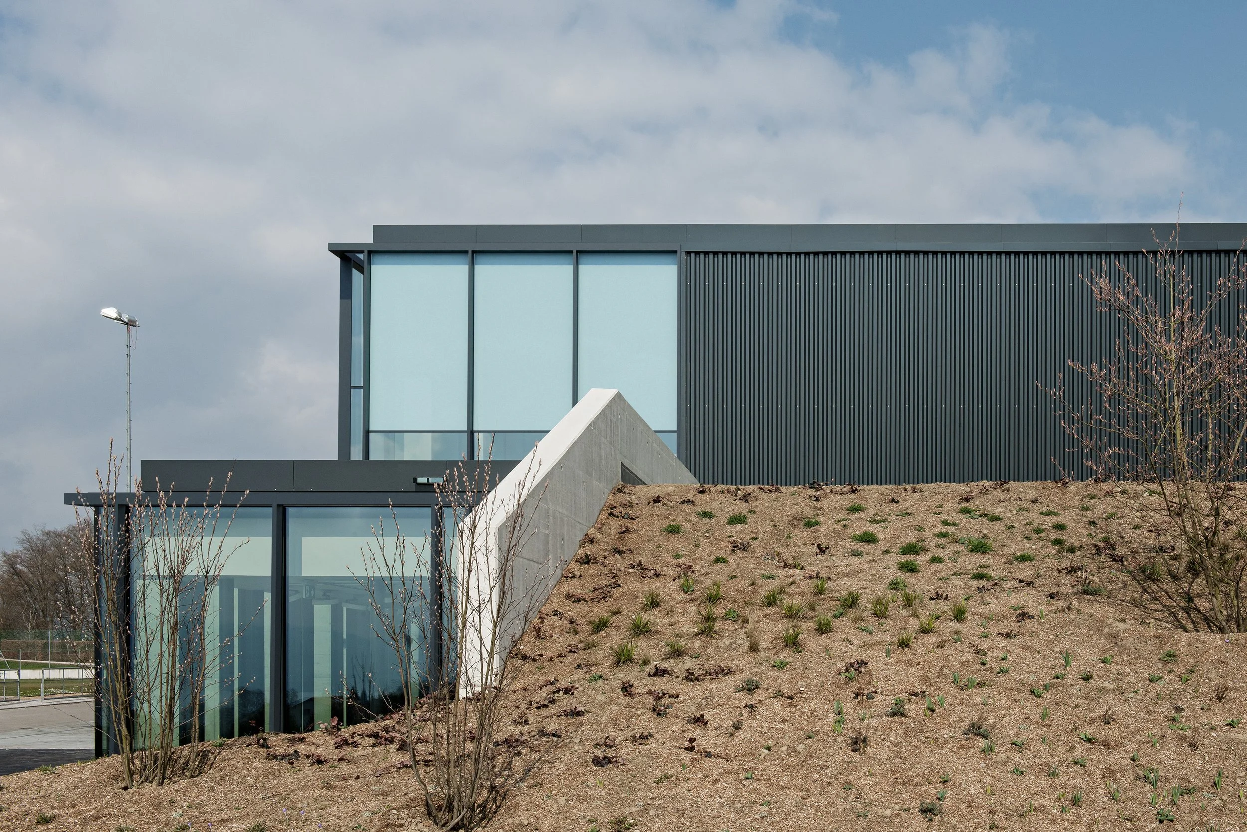 Modern building with large glass windows and black metal panels, situated on a sloped landscape with sparse plant growth and small trees, under a partly cloudy sky.