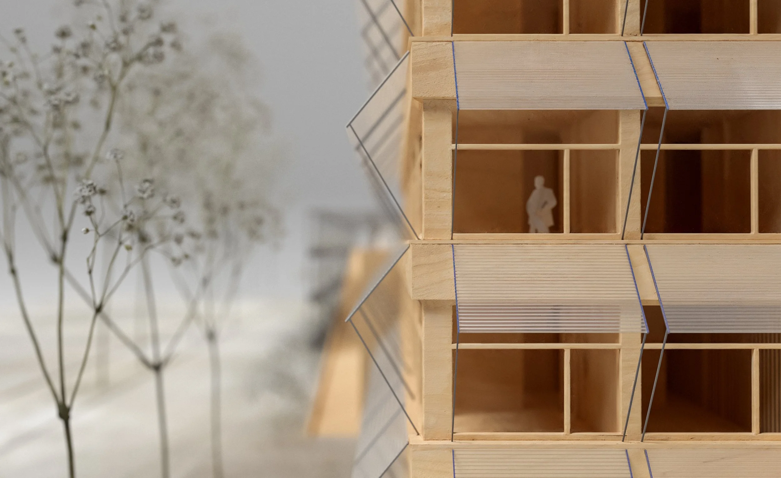 Architectural model of a building's interior and exterior, showing multiple floors and a tiny human figure inside for scale, with an abstract blurred floral element in the foreground.