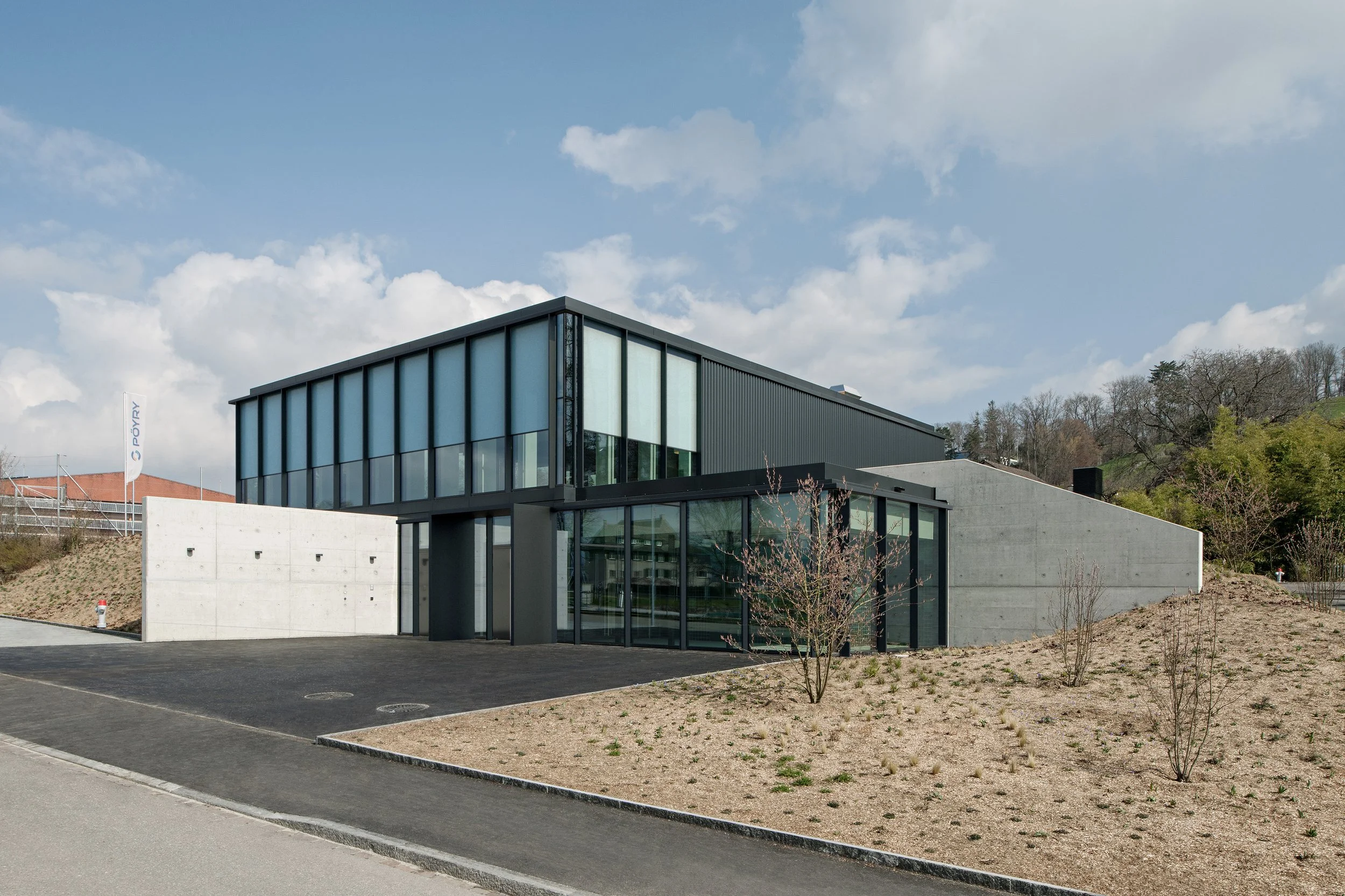 Modern building with black metal and large glass windows, surrounded by a dirt landscape with small plants, under a partly cloudy sky.