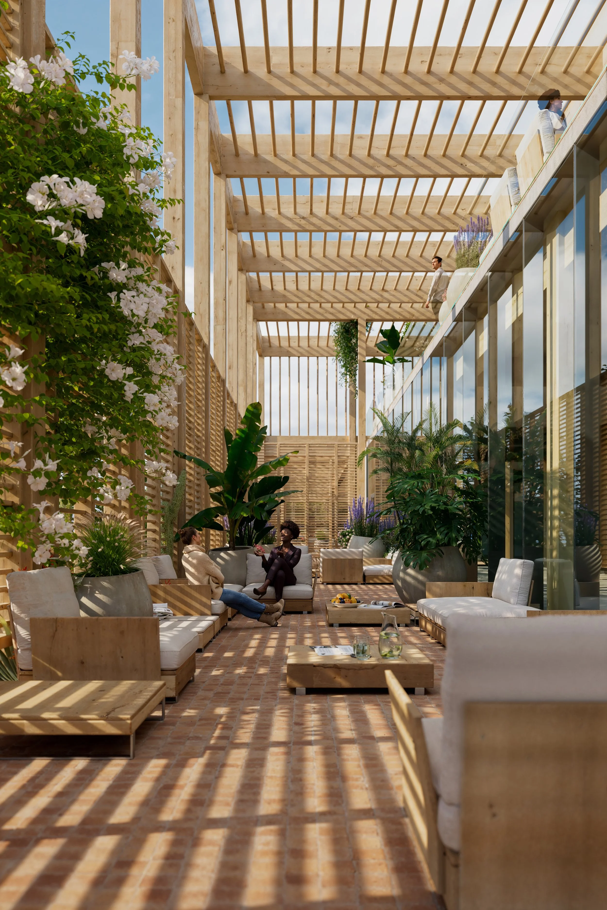 A modern outdoor patio area with wooden furniture, large potted plants, and a brick floor, with people sitting and chatting, surrounded by a wooden pergola structure and glass walls.
