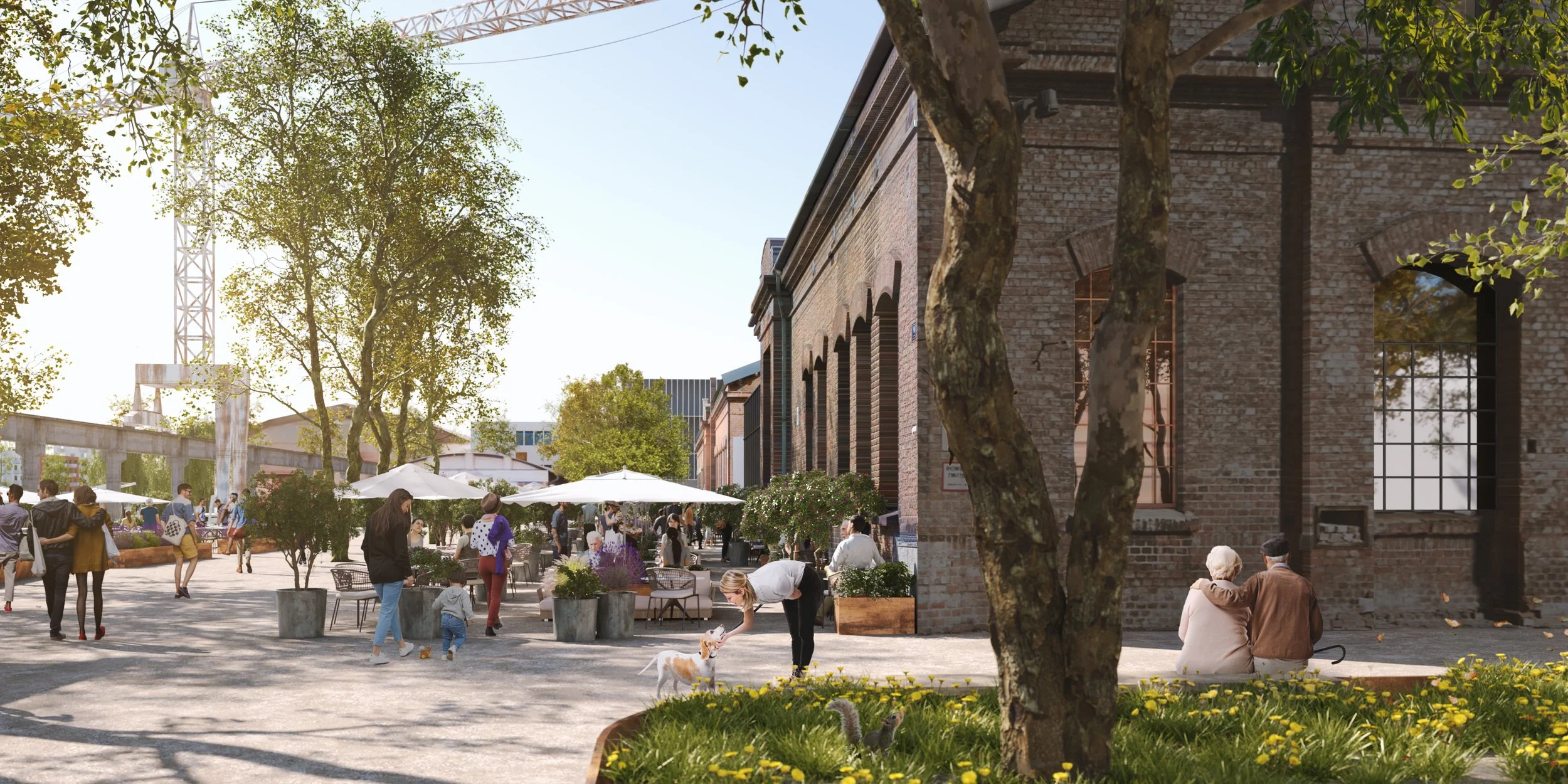 A lively outdoor urban scene with people walking and socializing on a sunny day, some sitting on a bench near a historic brick building, and a woman petting a small dog. There are large planters with flowers and trees, white umbrellas over tables, an