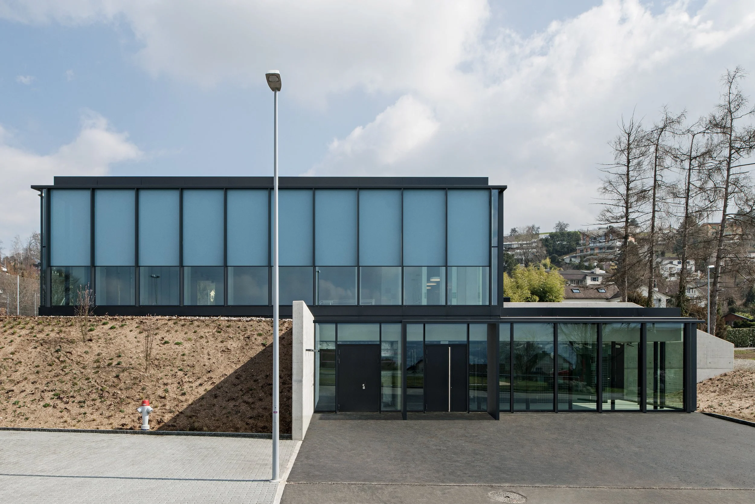 A modern two-story building with large blue-tinted glass windows, black framing, and a flat roof. The building is situated on a slightly sloped area with bare soil and a small garden patch. There is a streetlamp in the foreground and a fire hydrant n