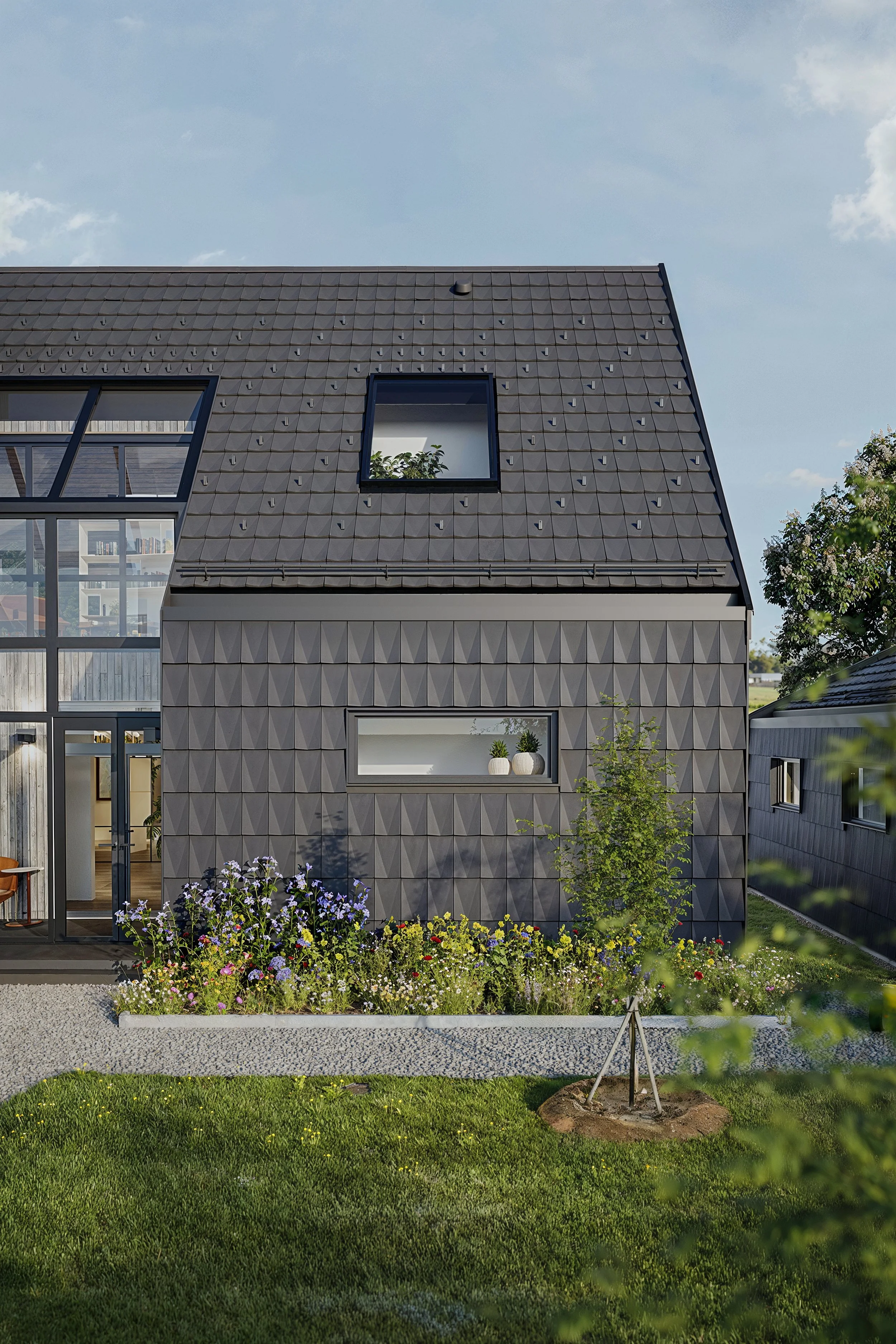 Modern house with dark gray textured exterior, flower bed with colorful flowers, green lawn, and a small tree, under a partly cloudy sky.