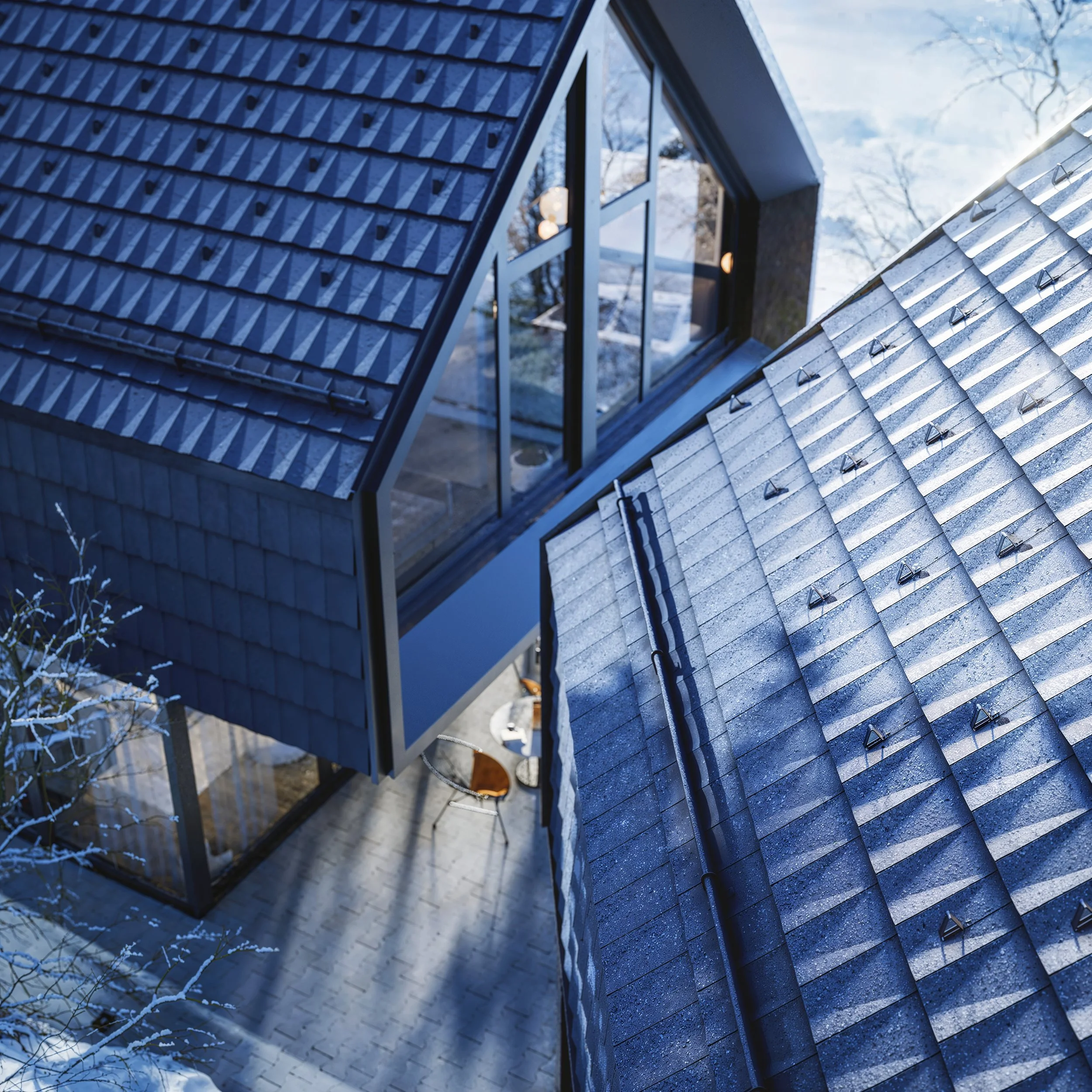 An aerial view of a modern house with a steep, dark blue tiled roof and large glass windows, surrounded by snow-covered trees.