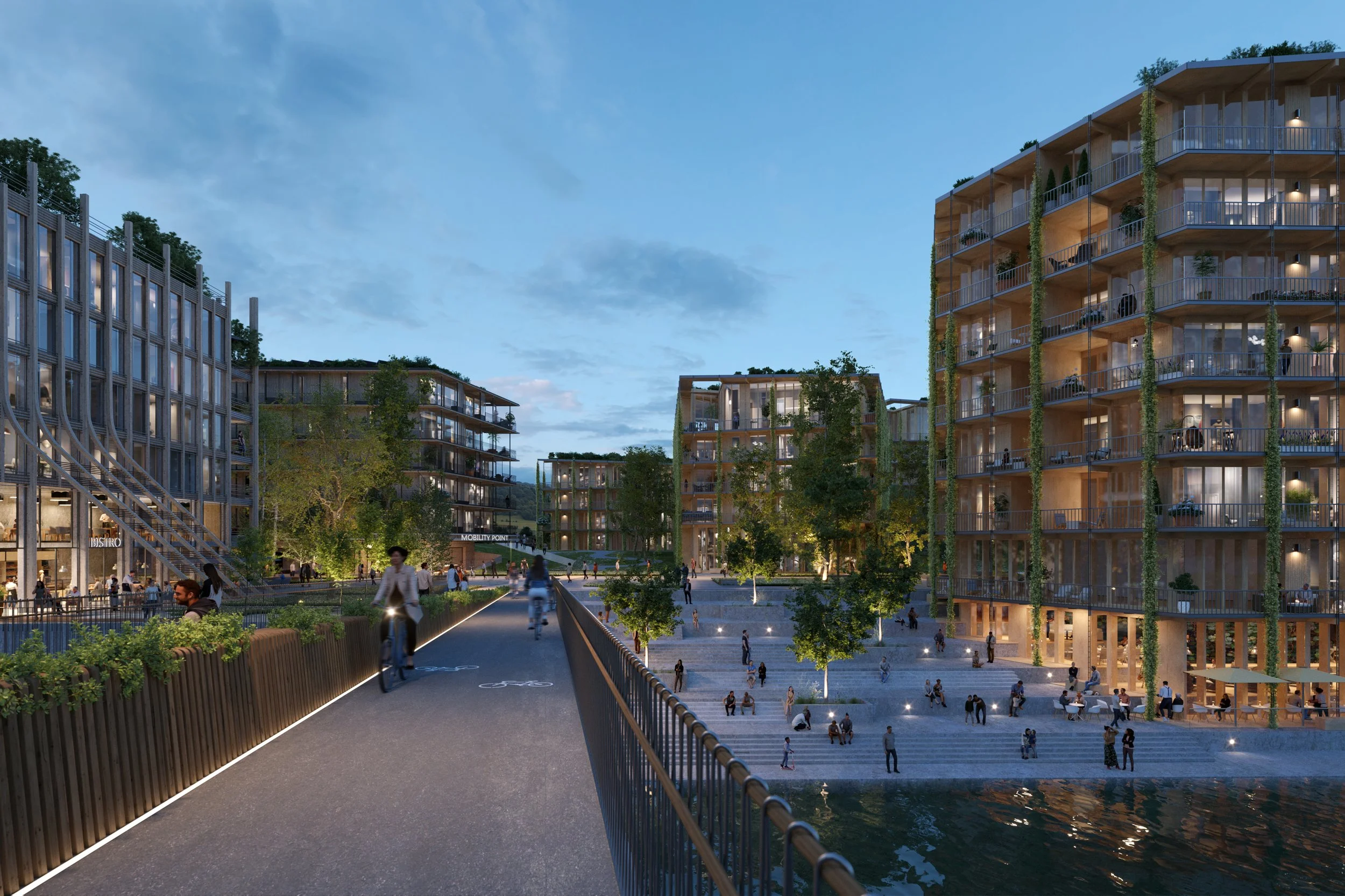 A modern residential complex with multiple multi-story buildings surrounding a waterfront and pedestrian area with trees, outdoor seating, and people walking, biking, and relaxing during dusk or early evening.