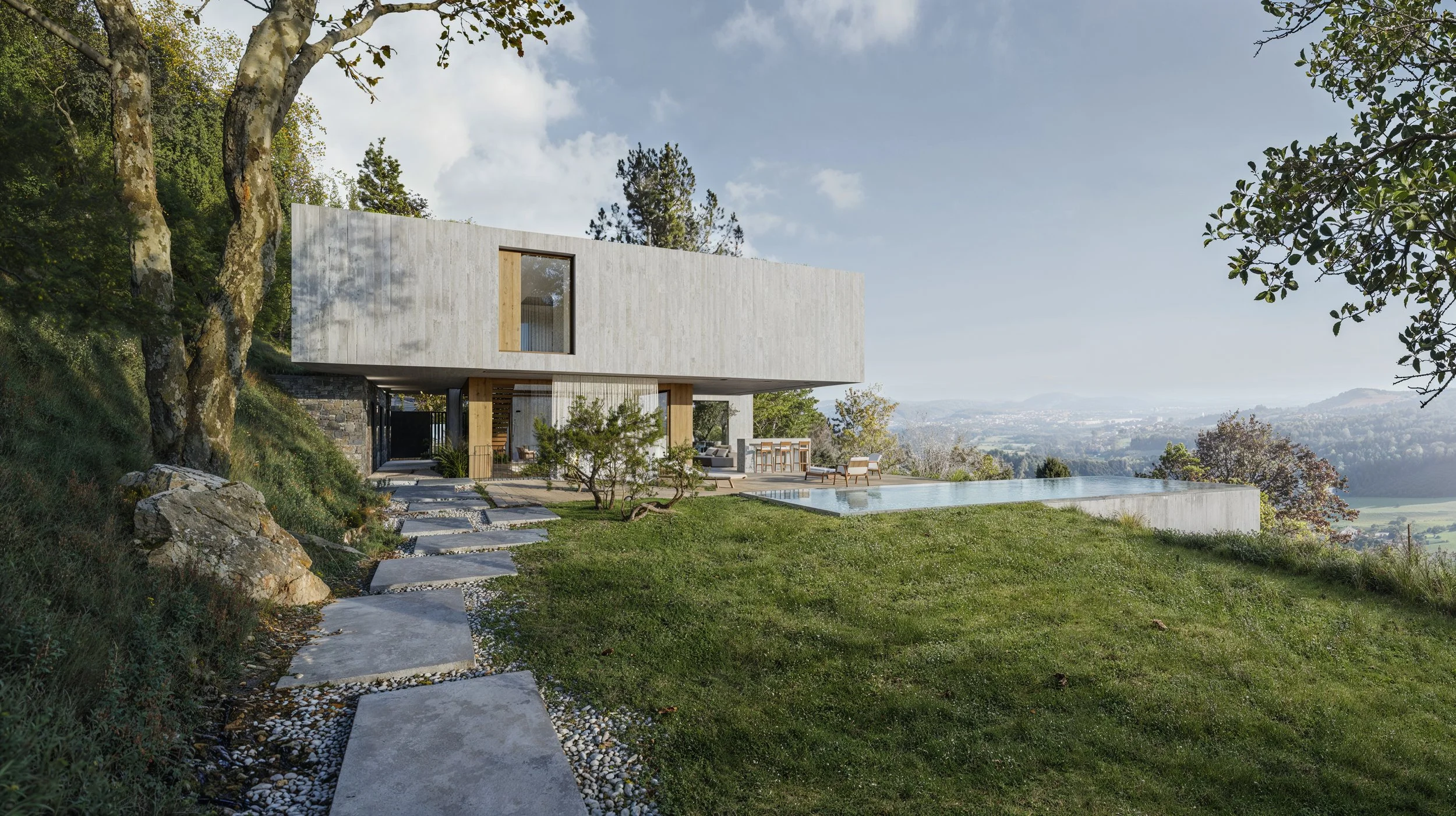 A modern house with a concrete exterior, large windows, and an outdoor patio area with furniture, set on a grassy hill with trees and a scenic view in the background.