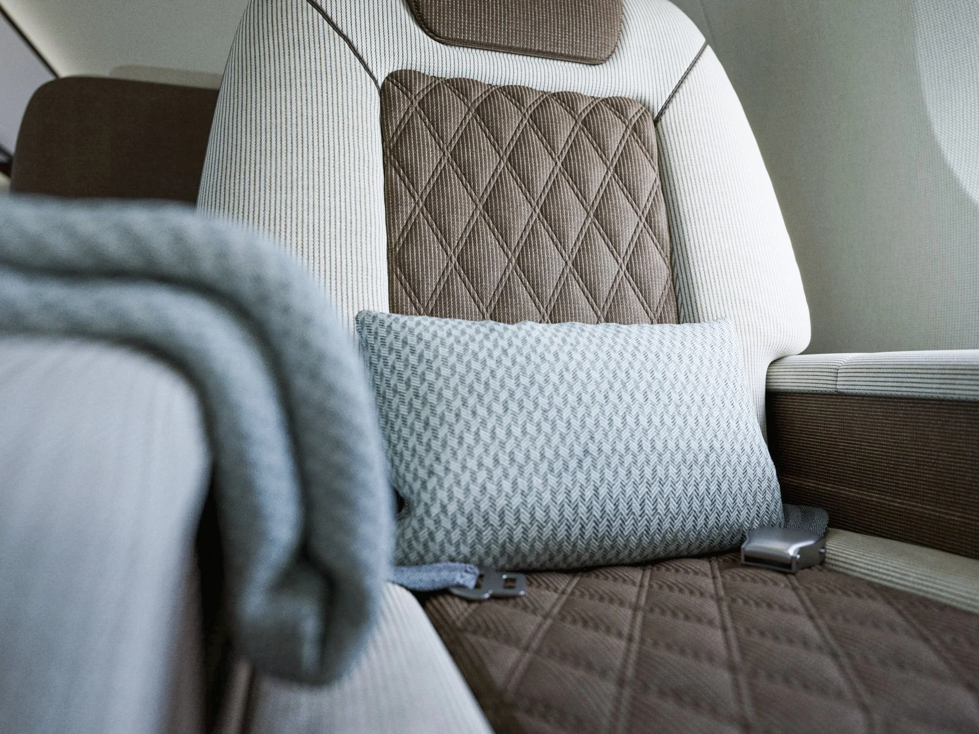 Close-up of a cushioned airplane seat with a pillow and blanket, featuring a diamond-patterned brown backrest and a beige striped wall in the background.