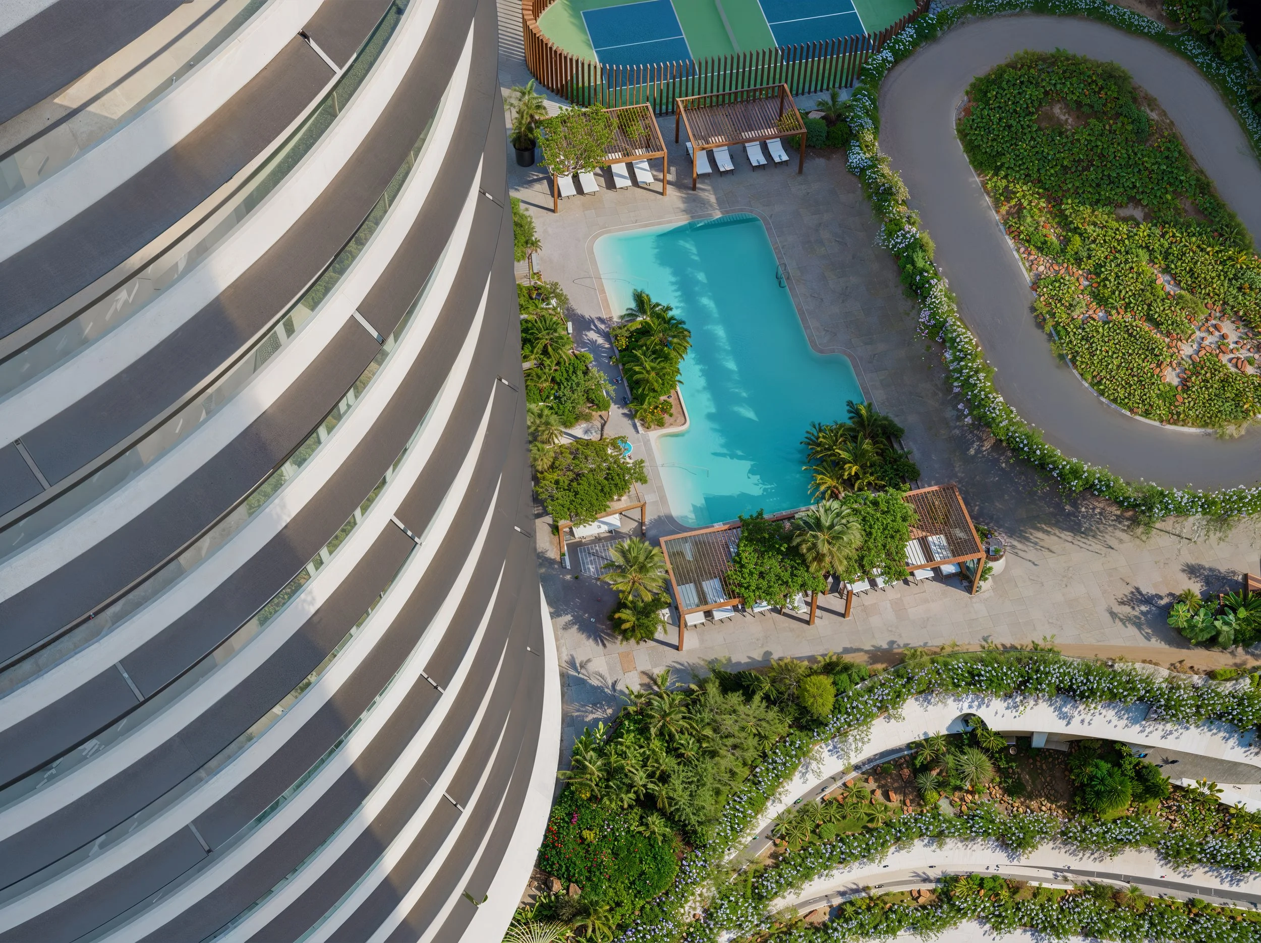 An aerial view of a hotel or apartment building with a curved glass facade, next to a swimming pool surrounded by lush greenery, lounge chairs, and shaded cabanas.