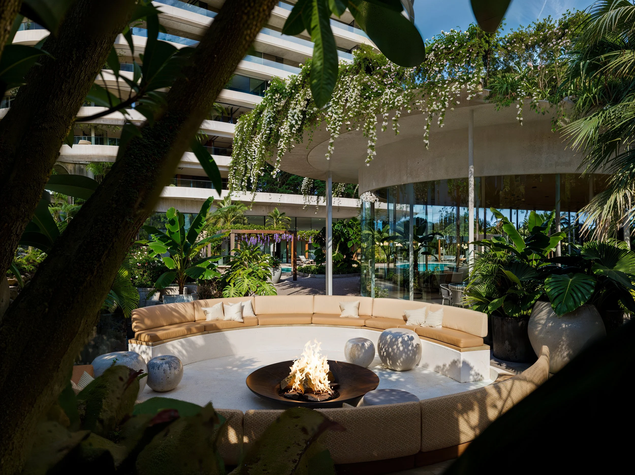 A modern outdoor lounge area with curved beige seating surrounding a small fire pit. The space is lush with tropical plants and trees, and a large glass building with balconies is visible in the background.