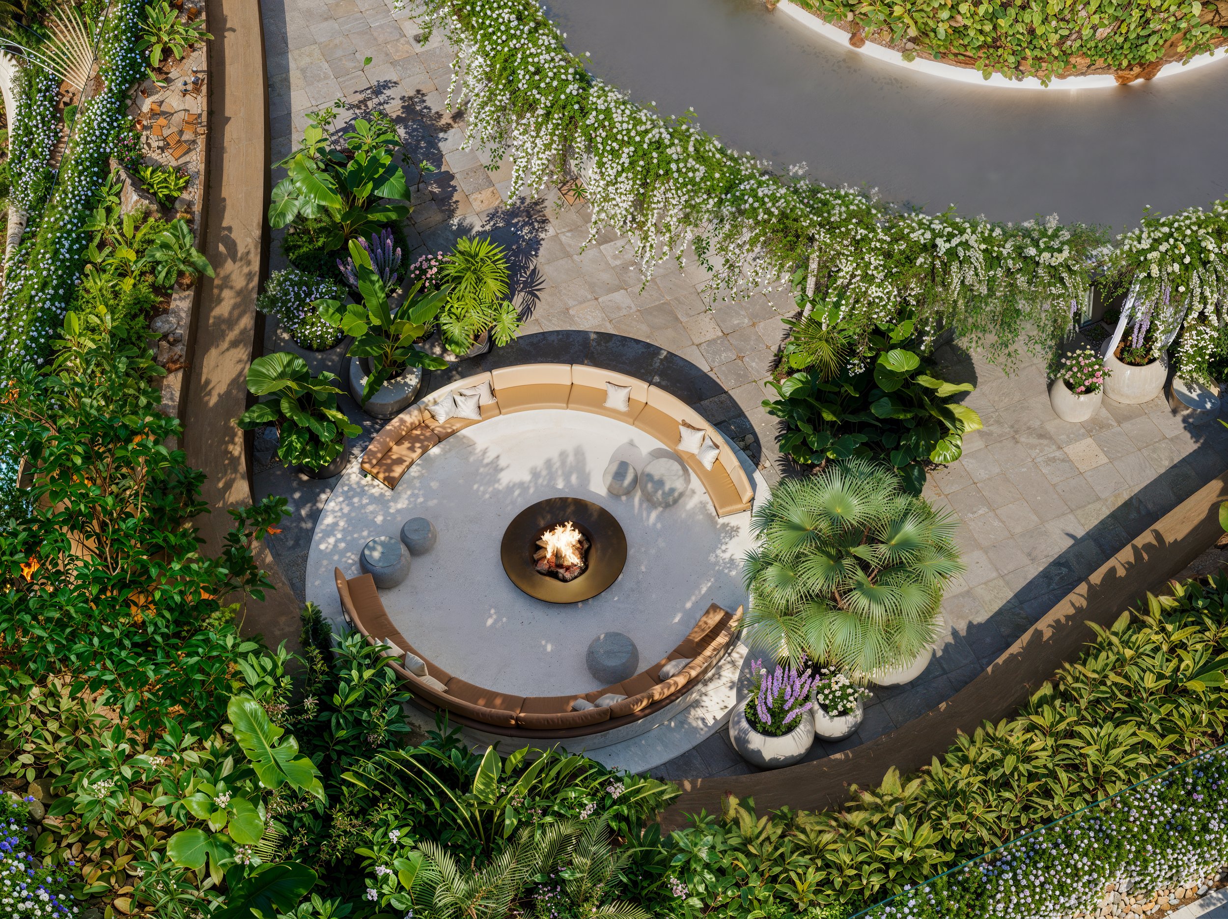 Aerial view of an outdoor garden seating area with a central fire pit, surrounded by a curved beige sofa, small round stools, and abundant lush green plants and flowering shrubs.