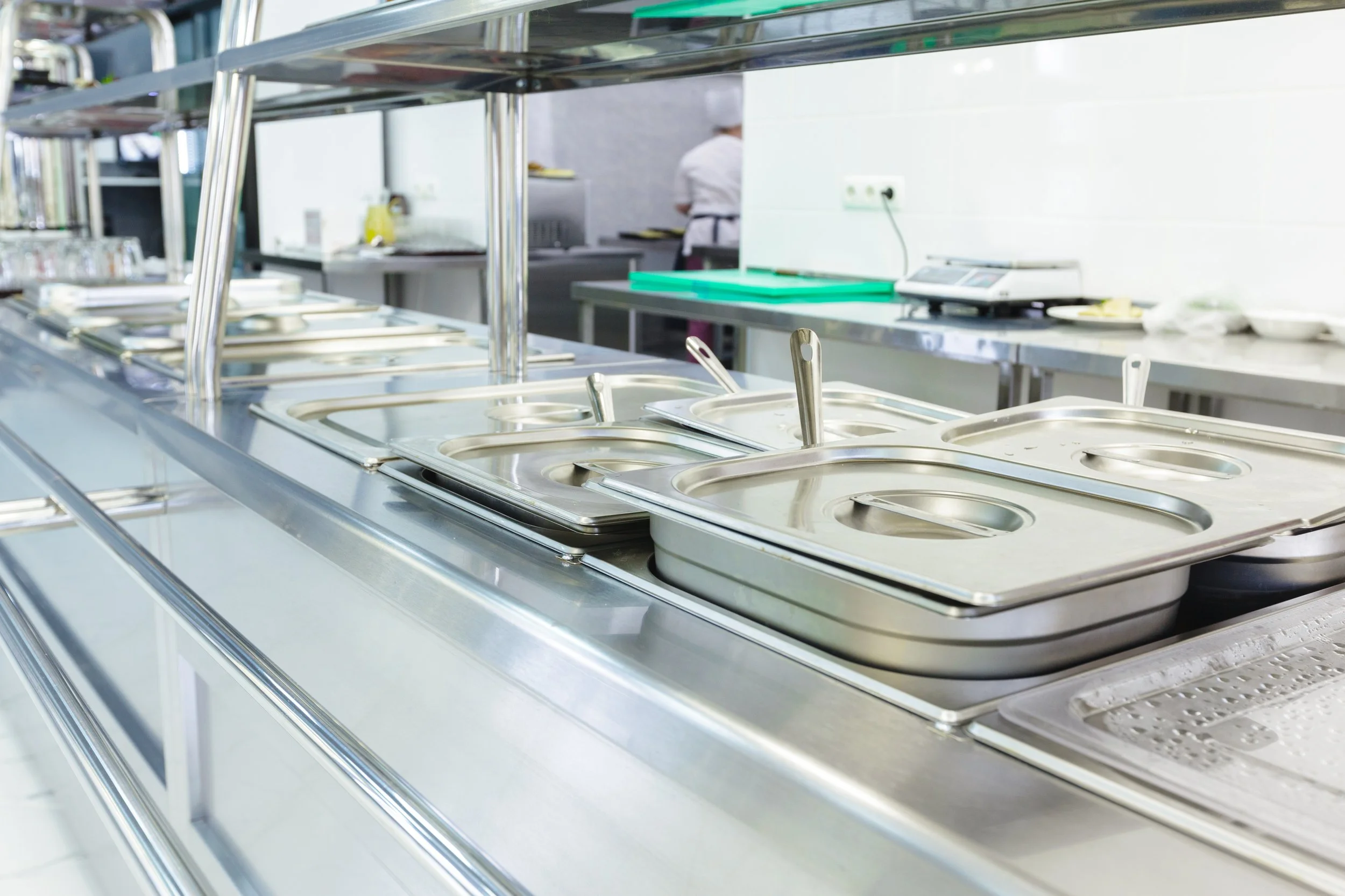 Empty stainless steel serving trays with lids on a cafeteria counter, with a chef working in the background.