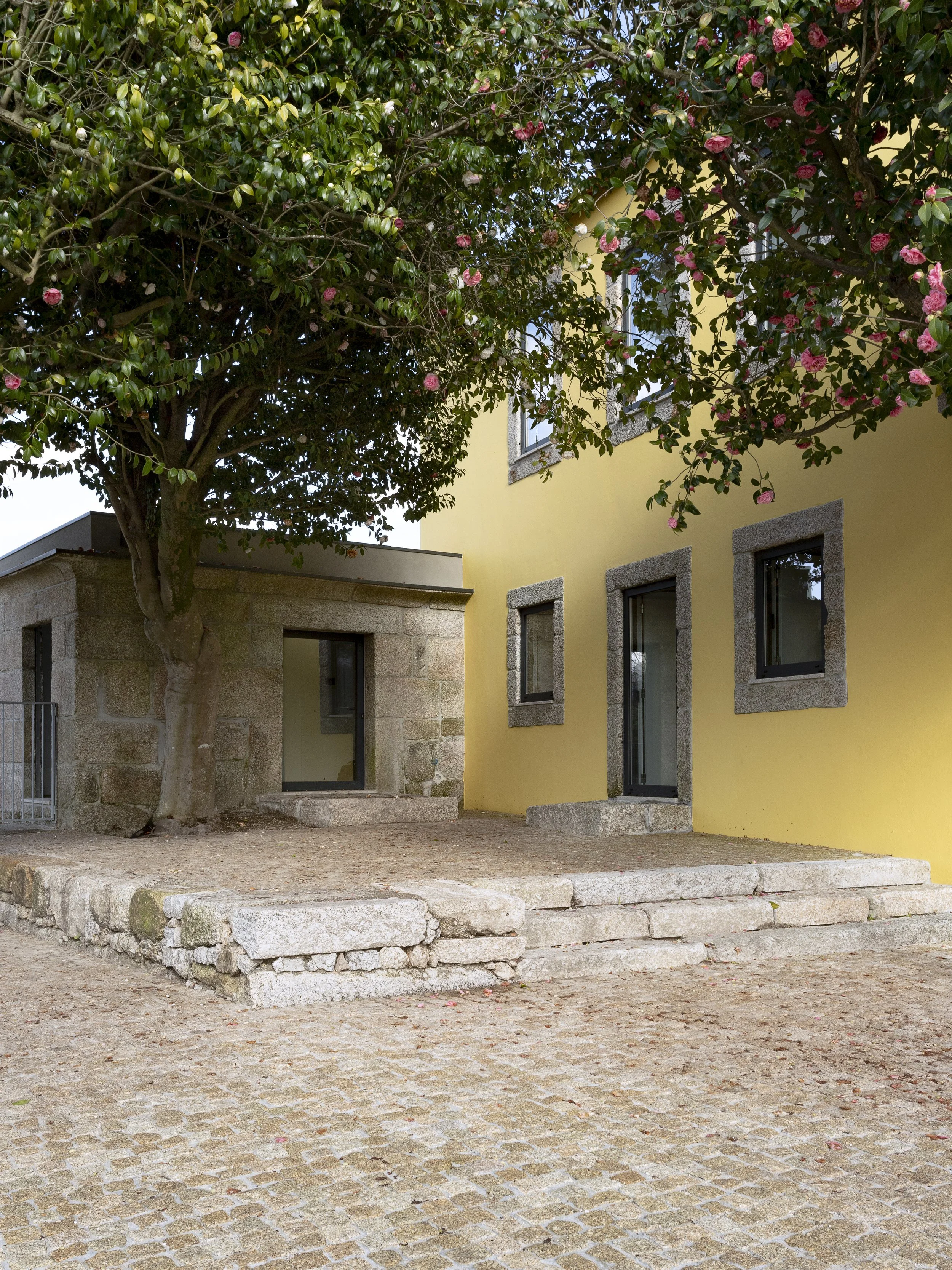 Edifício amarelo com janelas de pedra, acesso por escadas de pedra e uma árvore com flores rosas na frente.