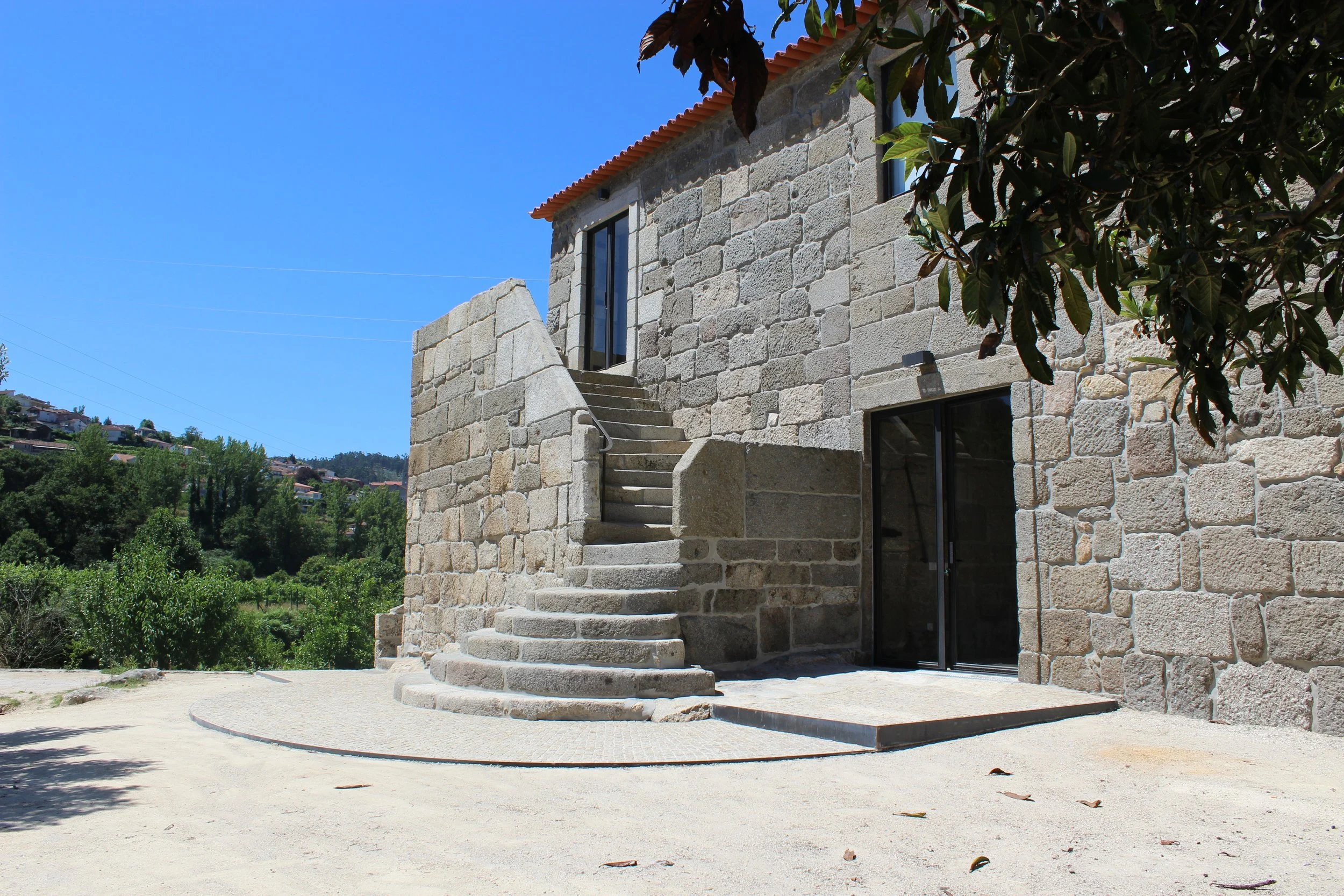 Prédio de pedra com escada curva de concreto na frente, janela de vidro e porta de vidro, cercado por árvores e céu azul.