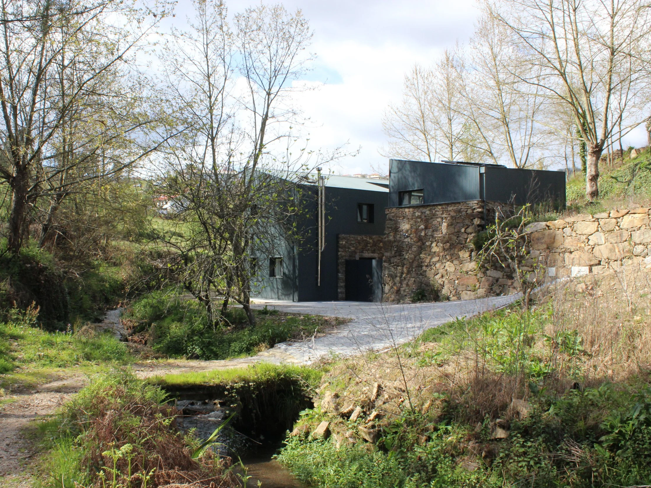 Casa moderna de parede de pedra e metal preto ao lado de riacho e árvores.
