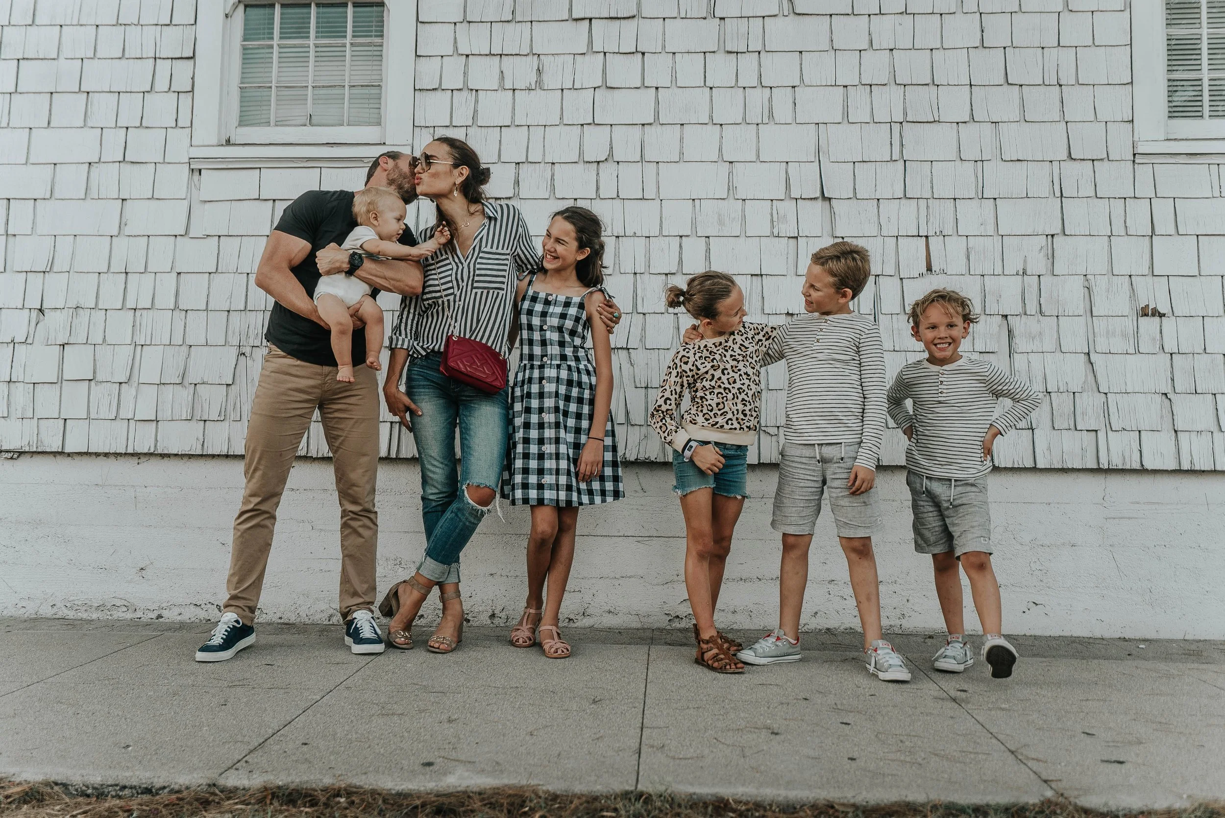 A family of seven standing outside against a white wooden wall. The parents are kissing, and the children are smiling and talking with each other.