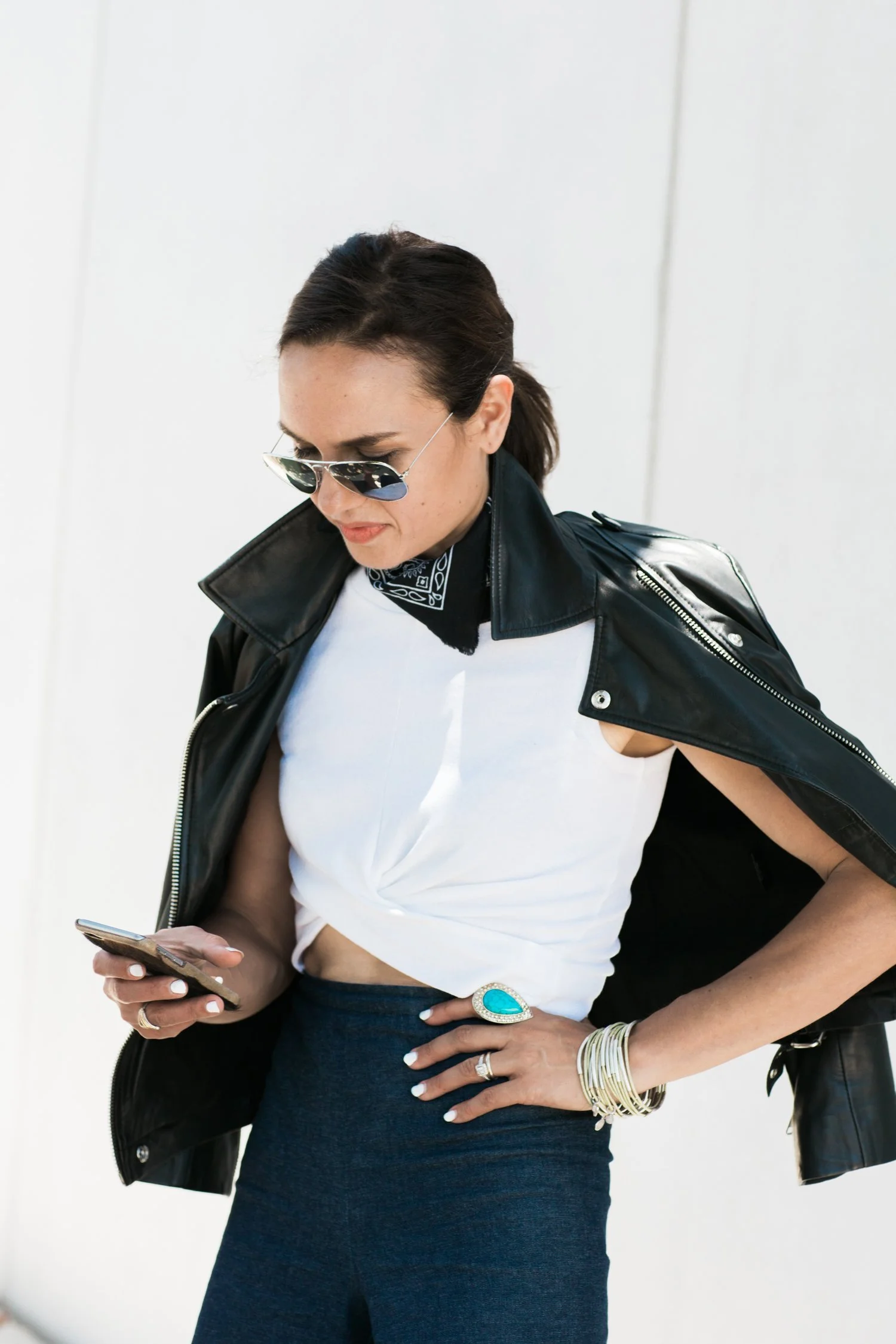 A woman wearing sunglasses, a black leather jacket draped over her shoulders, a white sleeveless top, blue high-waisted pants, and various jewelry including rings and bracelets, looking at her phone.
