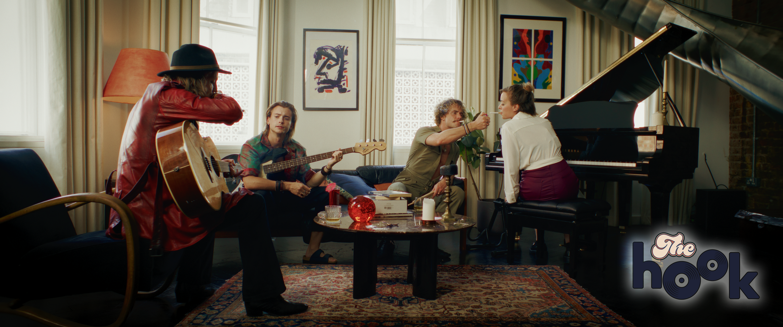 A group of five people in a cozy living room, some playing musical instruments and others singing, with colorful artwork on the walls and a grand piano. The image features a woman in a white shirt and burgundy skirt sitting at the piano, a man with curly hair and a beige jacket, a woman with a bass guitar, a person in a red jacket and hat playing an acoustic guitar, and another person with long hair sitting on the sofa. The room is decorated with a patterned rug, candles, and decorative objects.