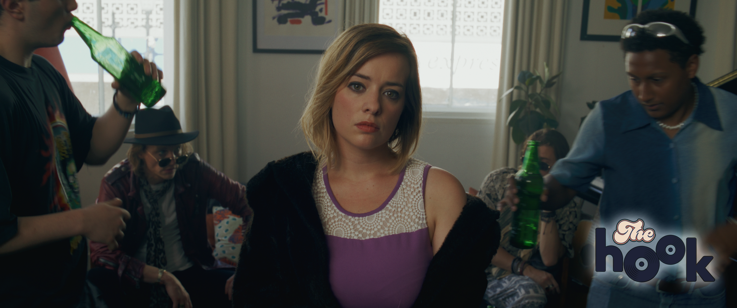 A woman with shoulder-length blonde hair and a serious expression sitting among several men and women at a party or social gathering in a room with a window, curtains, and artwork on the walls. Some people are holding green bottles, and one person is wearing sunglasses and a hat.