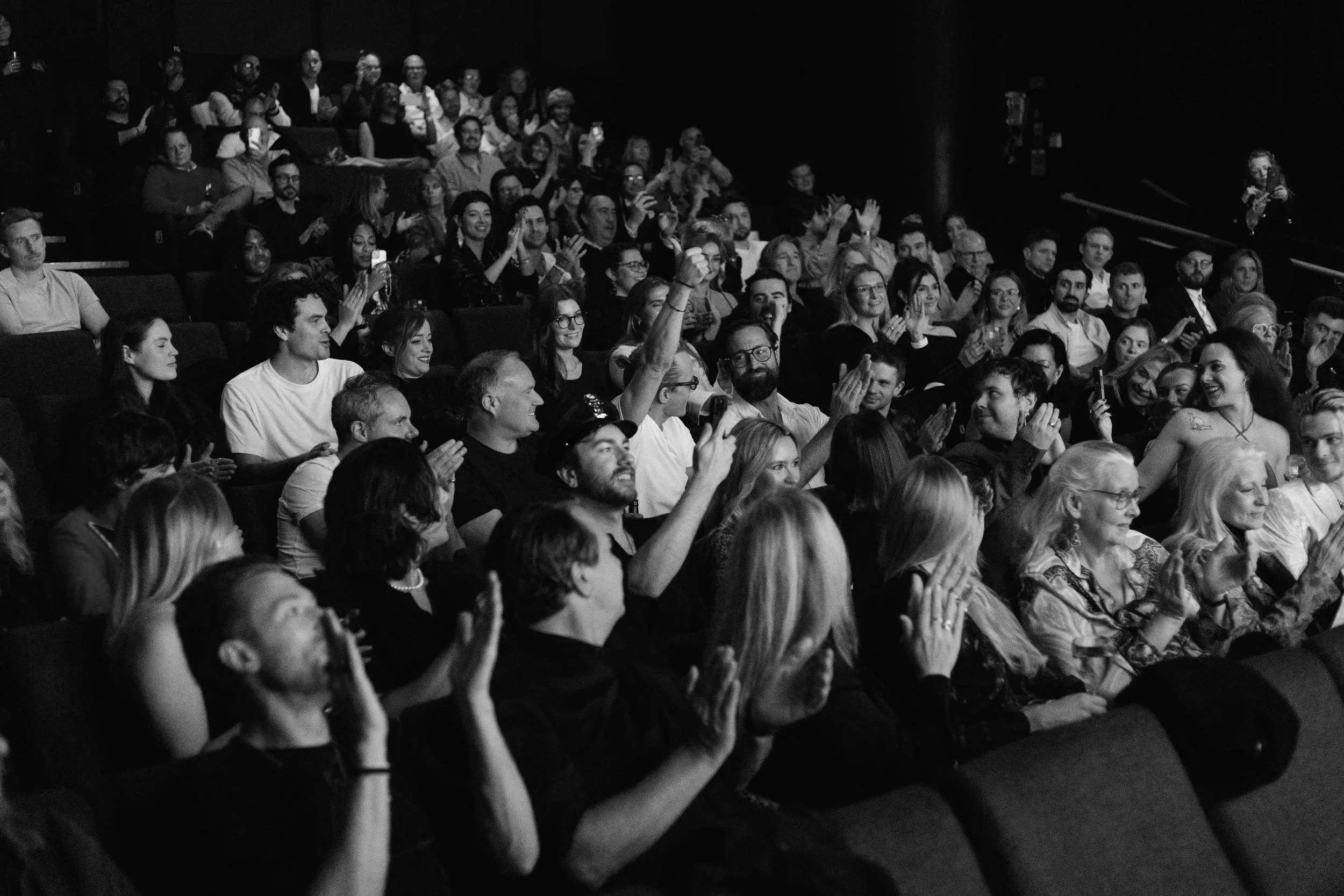 Black and white photo of an audience at a concert or event, clapping, smiling, and some raising their hands in admiration.