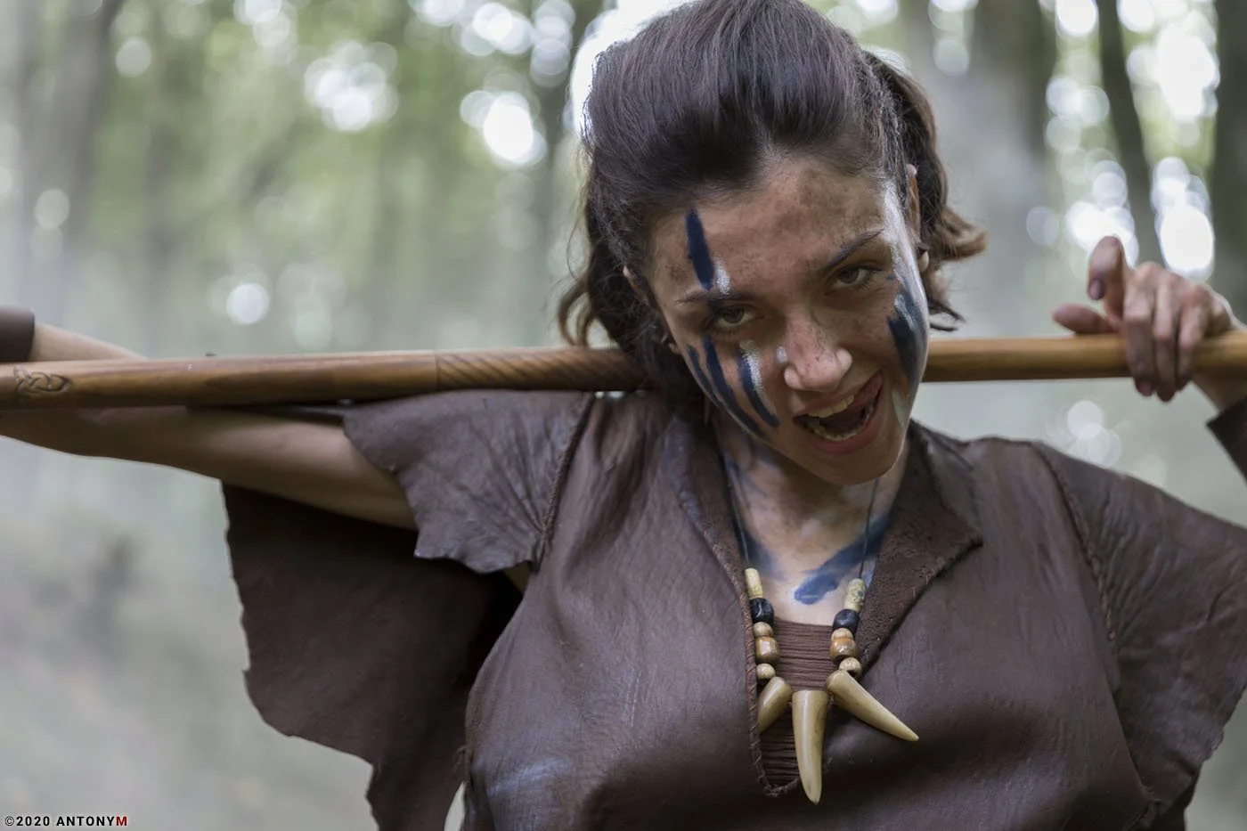 A woman with face paint and tribal jewelry carrying a wooden staff on her shoulders in a forest.