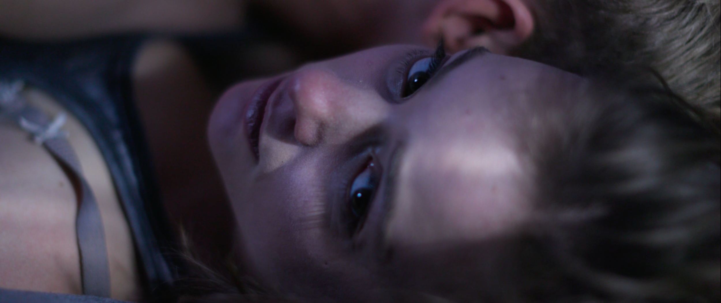 Close-up of a woman's face with expressive eyes, lying down with her head tilted, and a person's face partially visible in the background.