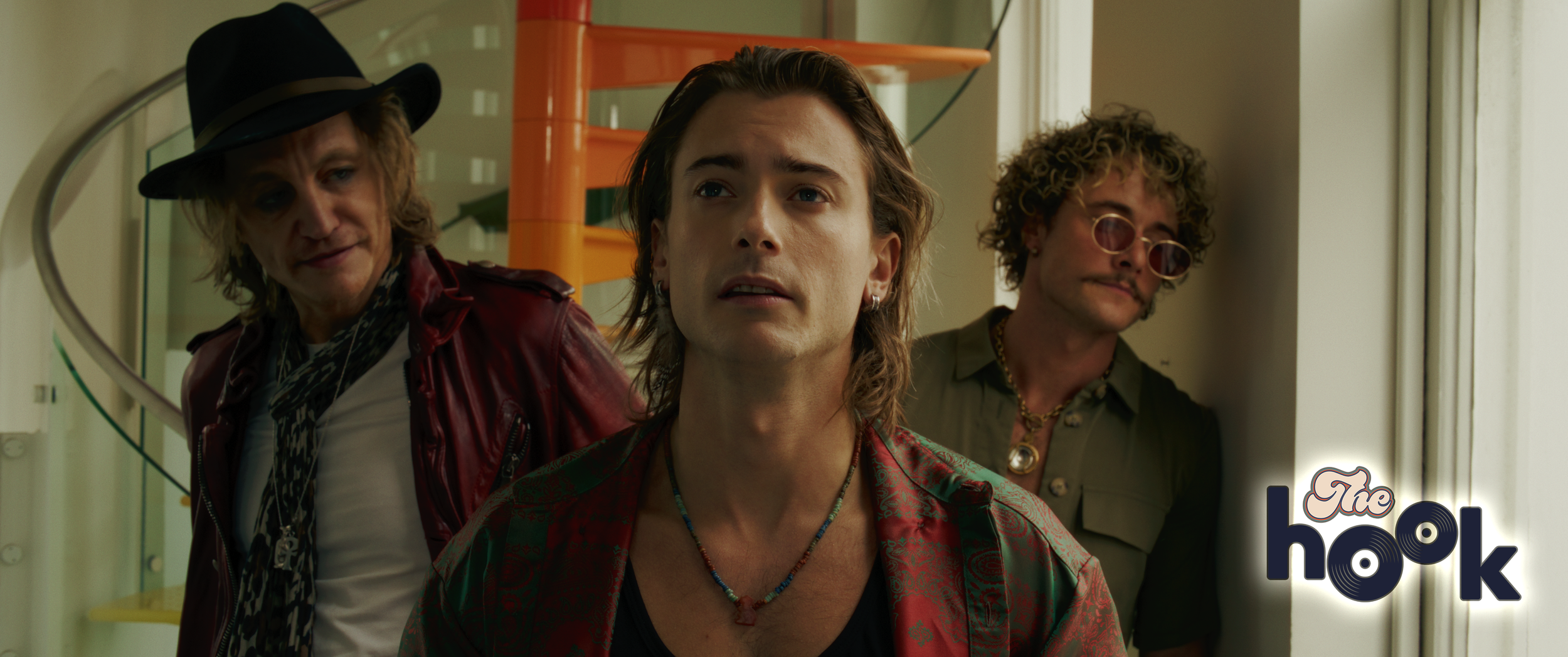 Three men standing inside a building, with one near a window, looking contemplative. The man on the left is wearing a black fedora, red leather jacket, and patterned shirt. The man in the center has long hair, earrings, and colorful necklaces. The man on the right has curly hair, glasses, and a green shirt. The logo of the film 'The Hook' is in the bottom right corner.