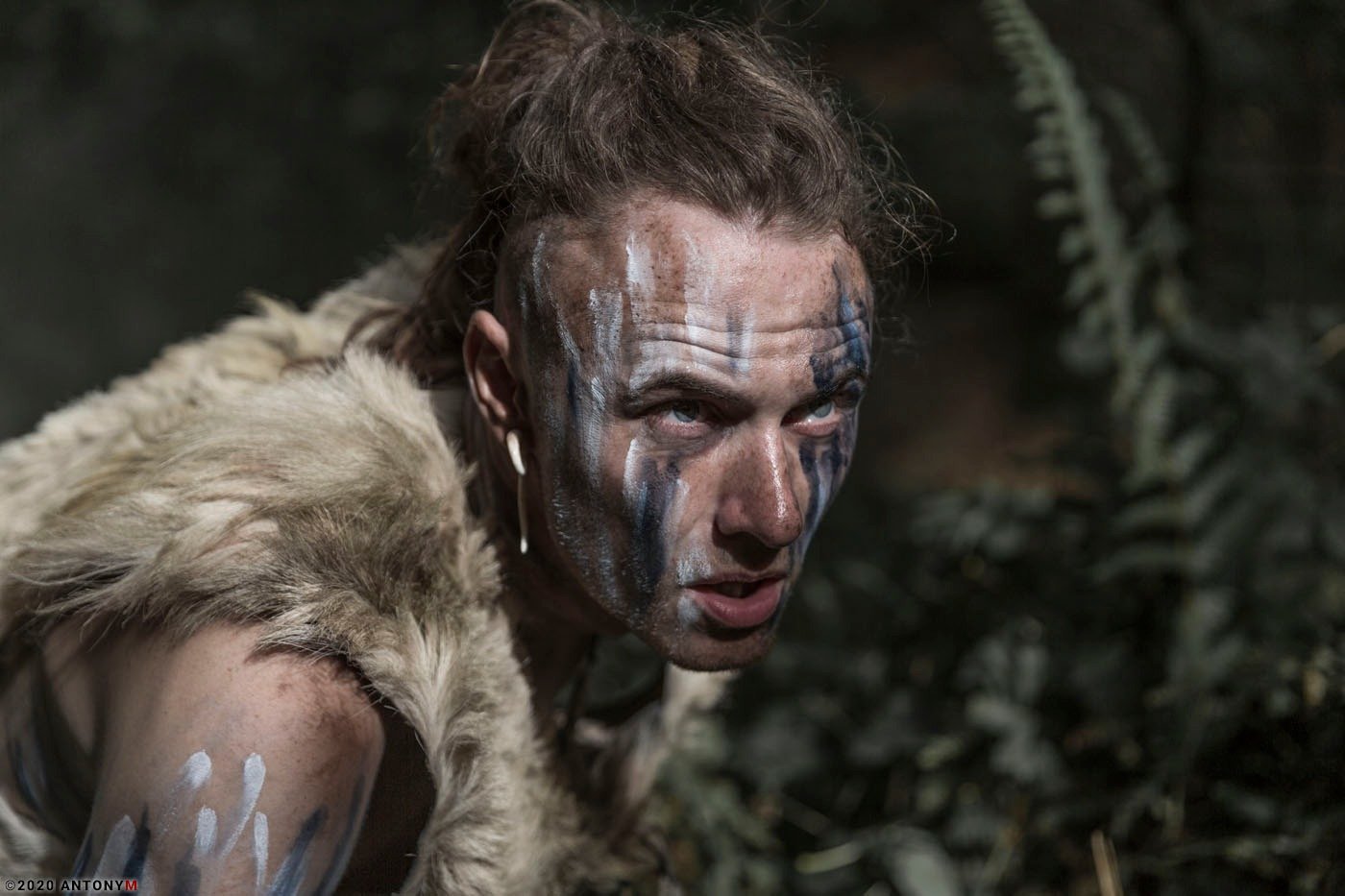 A woman with face paint and tribal clothing, posing outdoors in a forested area.