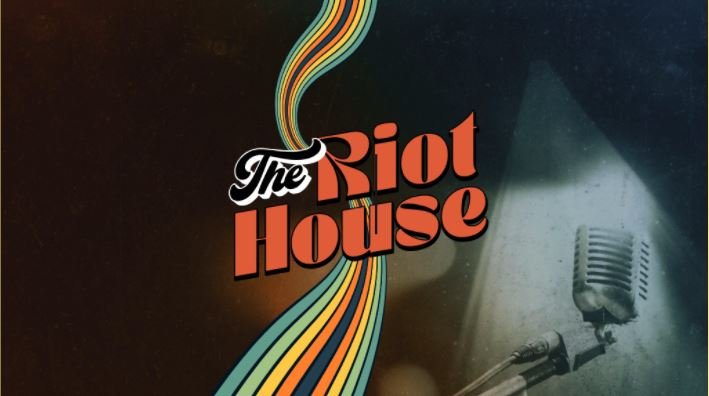 Close-up of a vintage microphone on a stand with a dark, smoky background. The image features a colorful, swirling rainbow-like design and the text 'The Riot House' in bold red and black font.