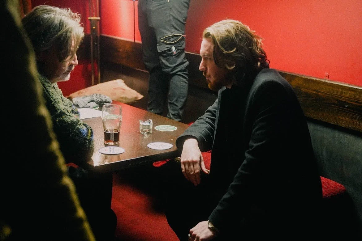 Two men are seated at a table in a dimly lit restaurant or bar, engaging in conversation. There are drinks on the table, including a glass of beer and a small glass of water. The background features a red wall with wooden accents.
