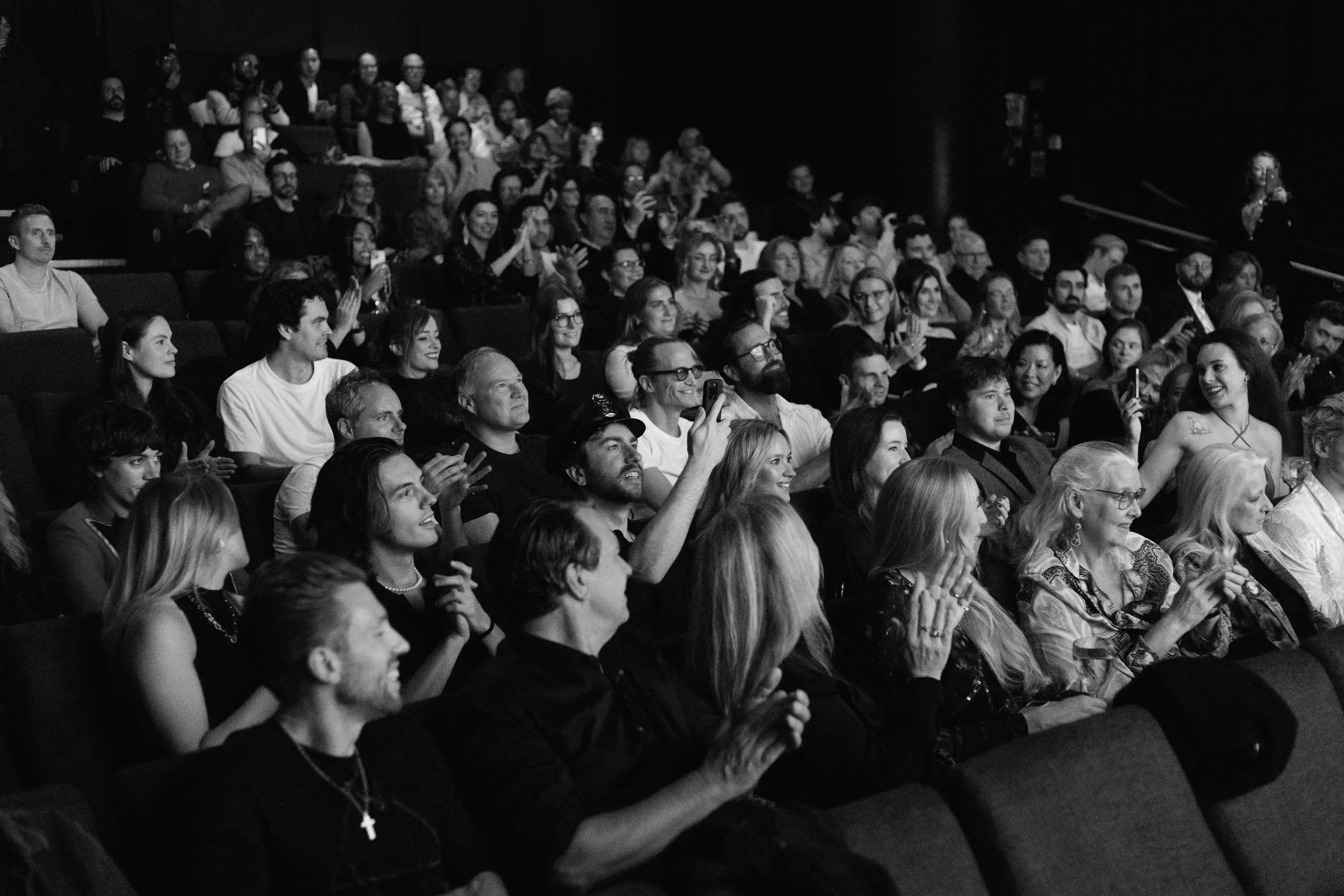 Audience watching a performance or event in a theater, clapping and smiling, black and white photo.
