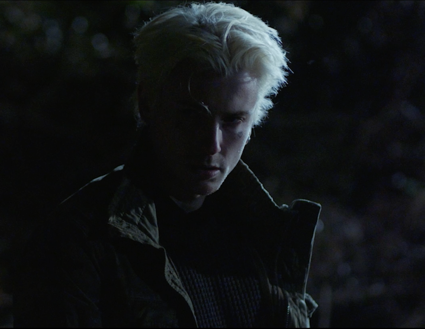 A person with platinum blonde hair illuminated in low light, with a dark background.