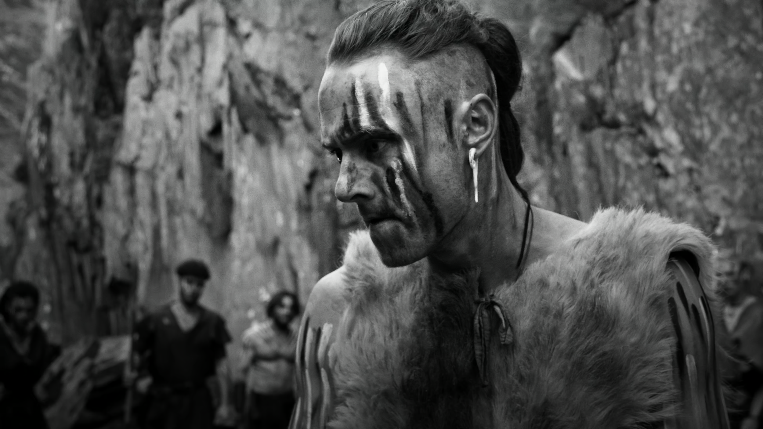 A person with face paint, wearing fur clothing and earrings, looks to the side. Four others stand in the background near a rocky landscape.