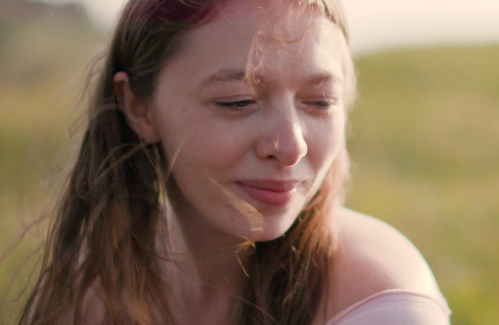 Close-up of a young woman with long brown hair outdoors, looking down with a gentle smile, in natural light.