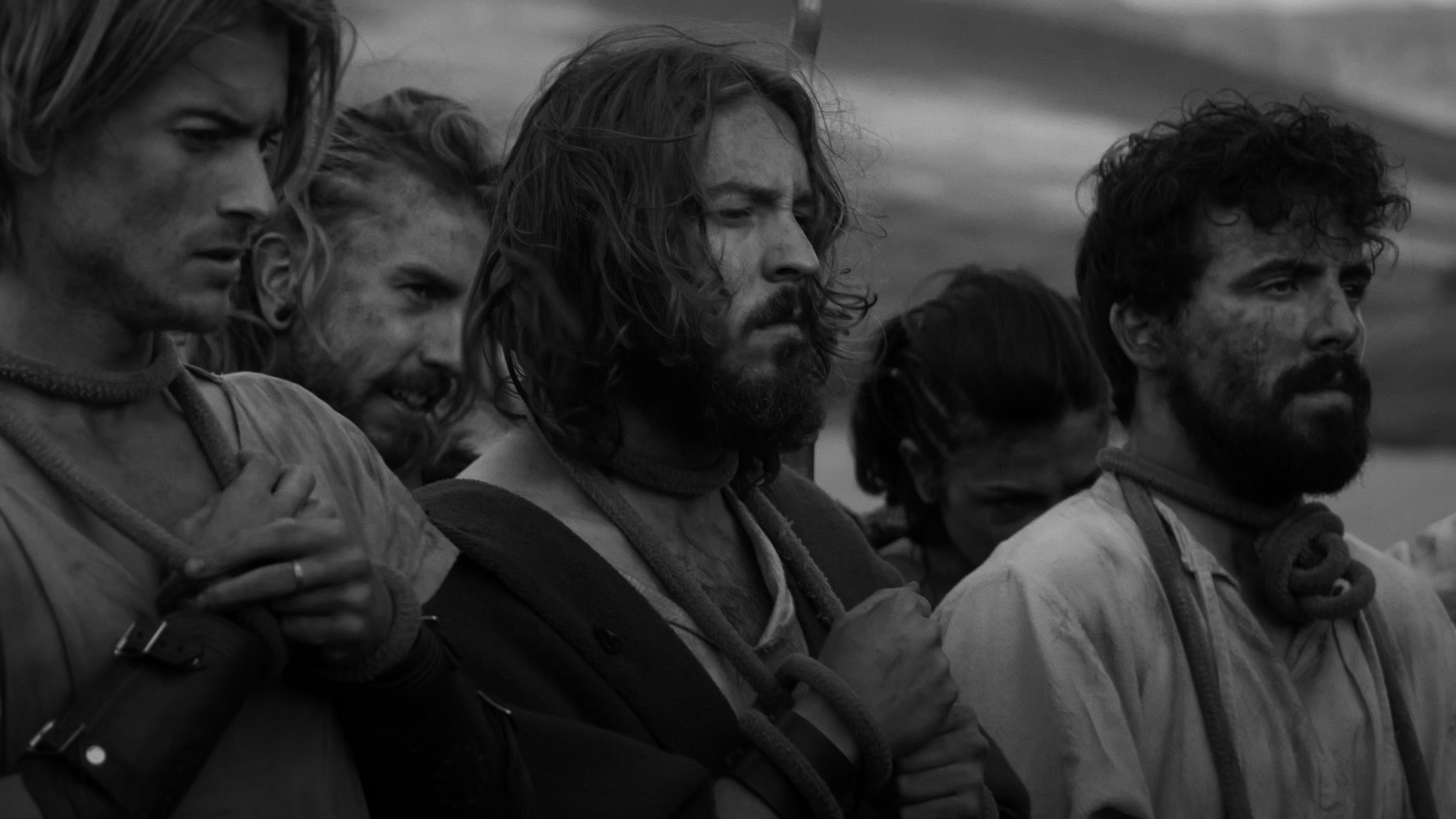 Close-up of five men with long hair and beards, standing in line with their hands on their chests, looking serious and solemn, wearing outdoor clothing, in black and white.
