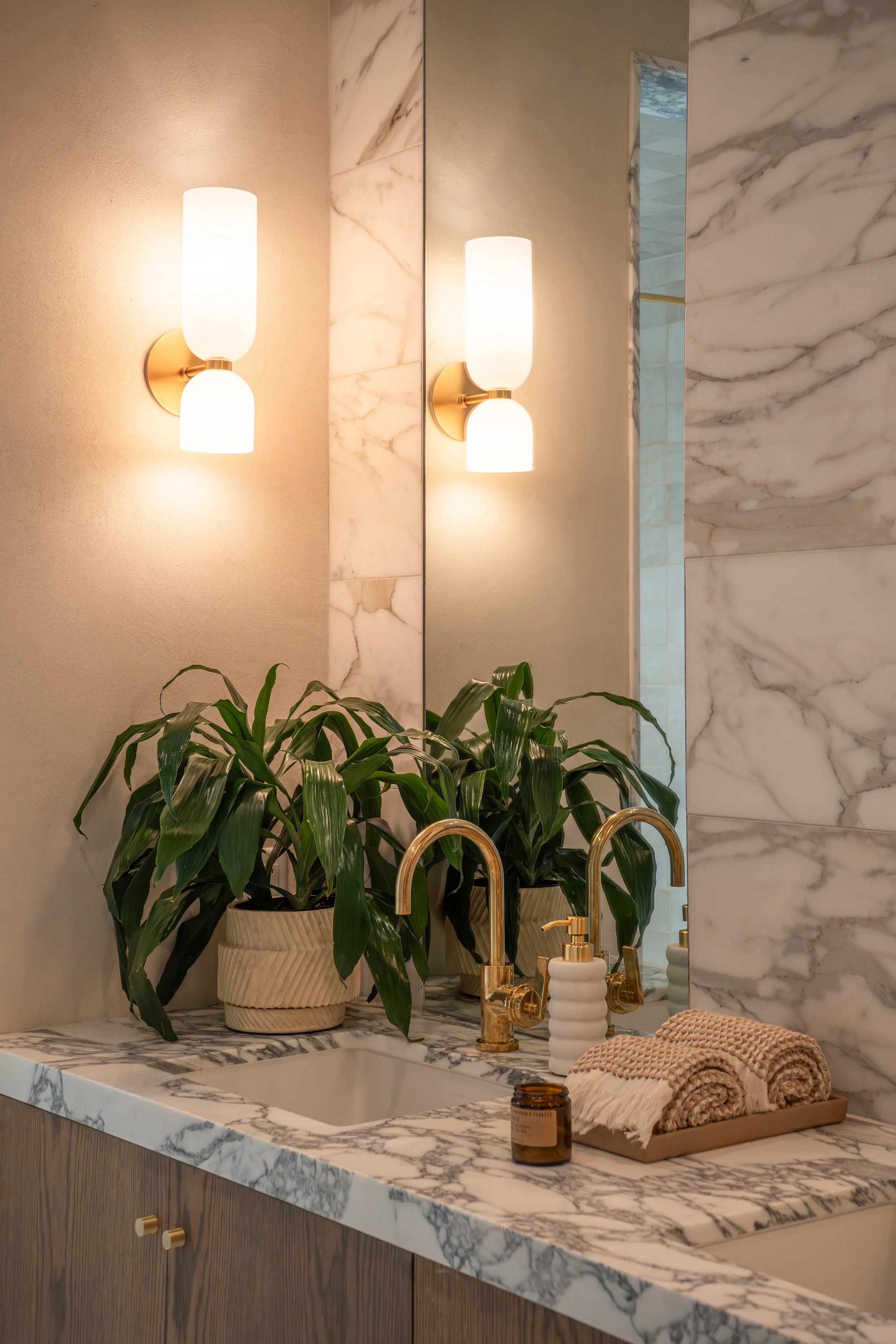 A bathroom vanity with a marble countertop, green potted plant, gold faucets, and a tray with rolled towels and a candle, with a mirror and wall-mounted lights