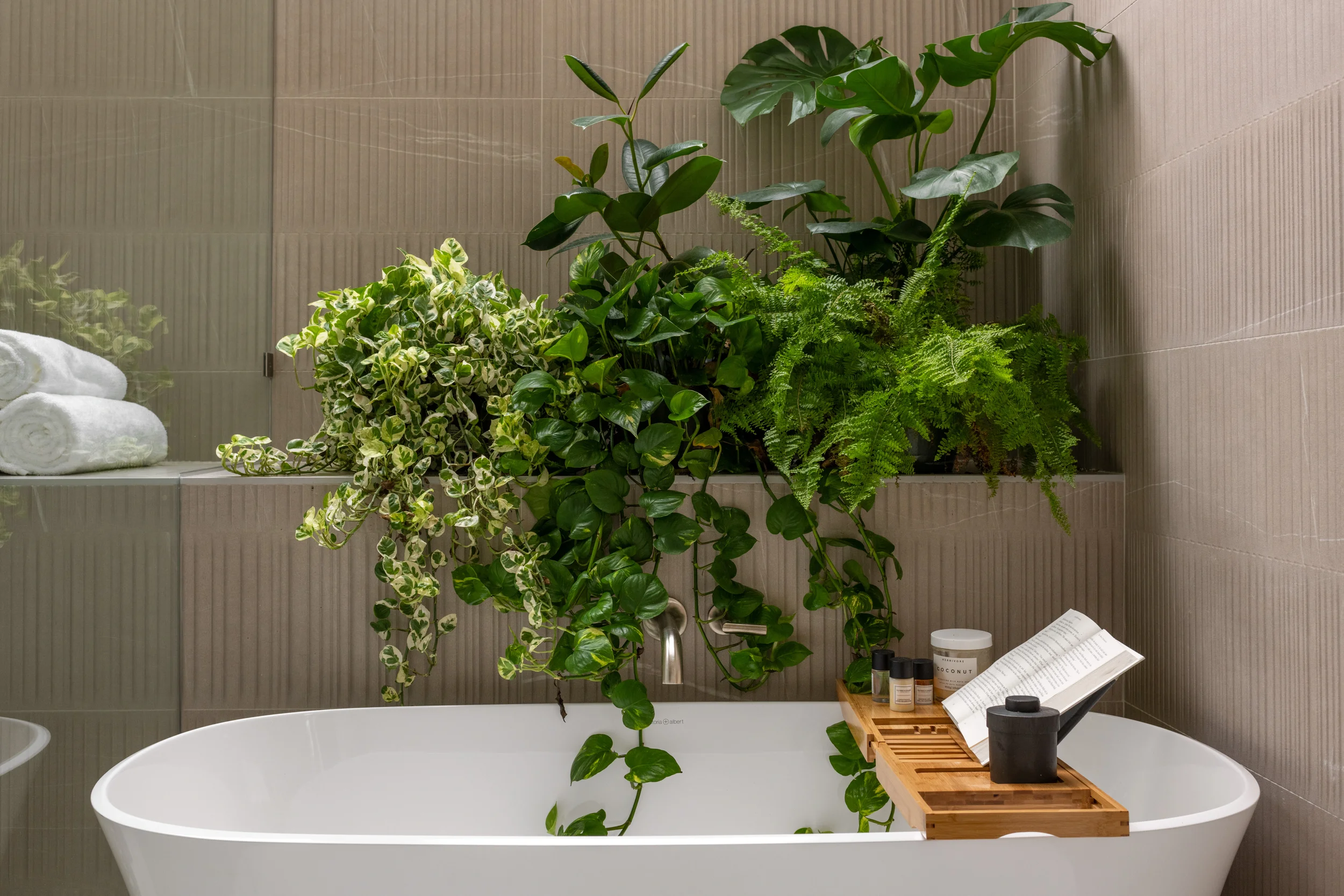 Bathroom with a white bathtub surrounded by green plants and towels, with a wooden bath tray holding toiletries and a book.