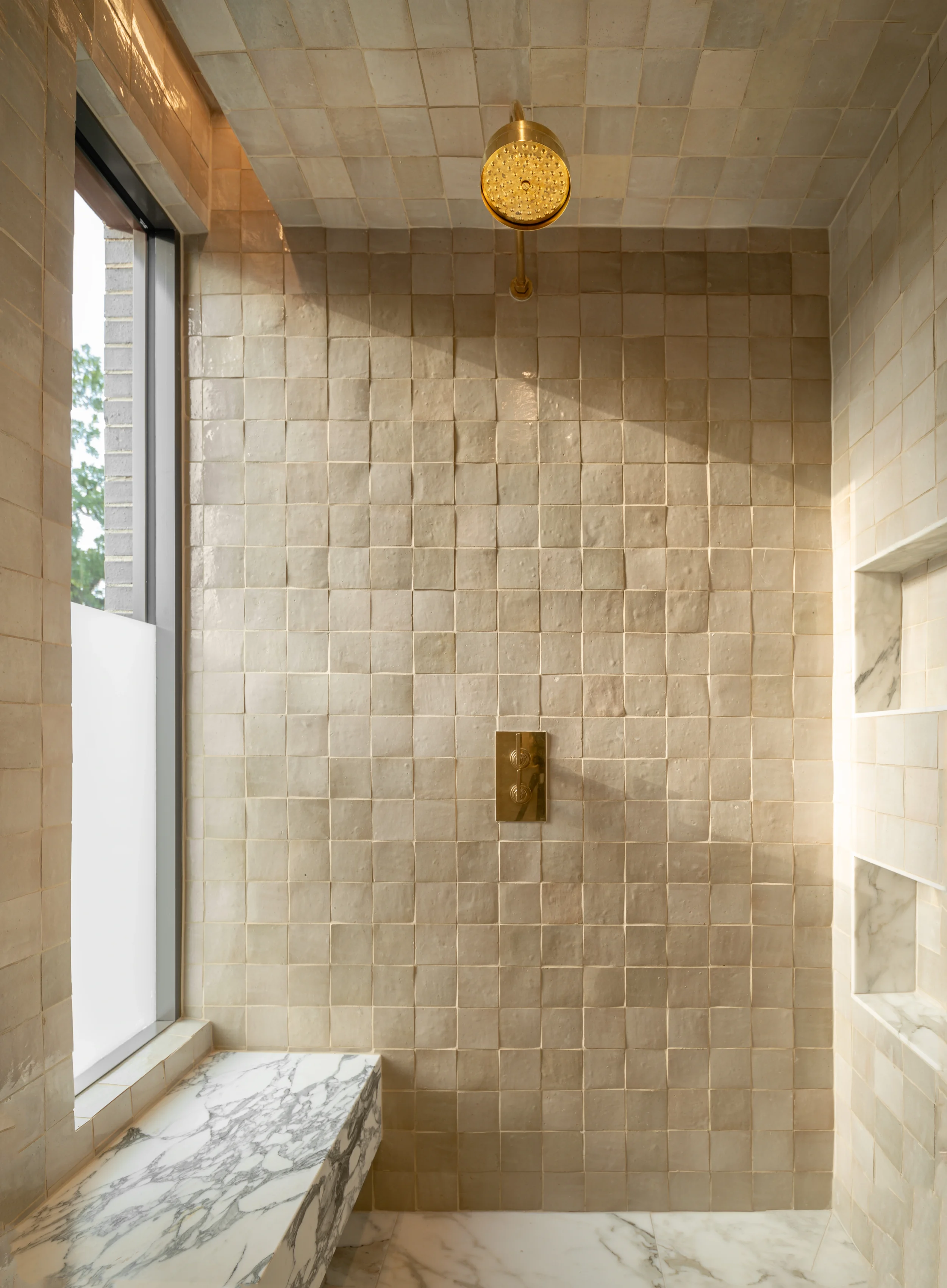 A luxurious shower with beige tile walls, a round gold showerhead, a built-in marble seat, and a window with frosted glass.