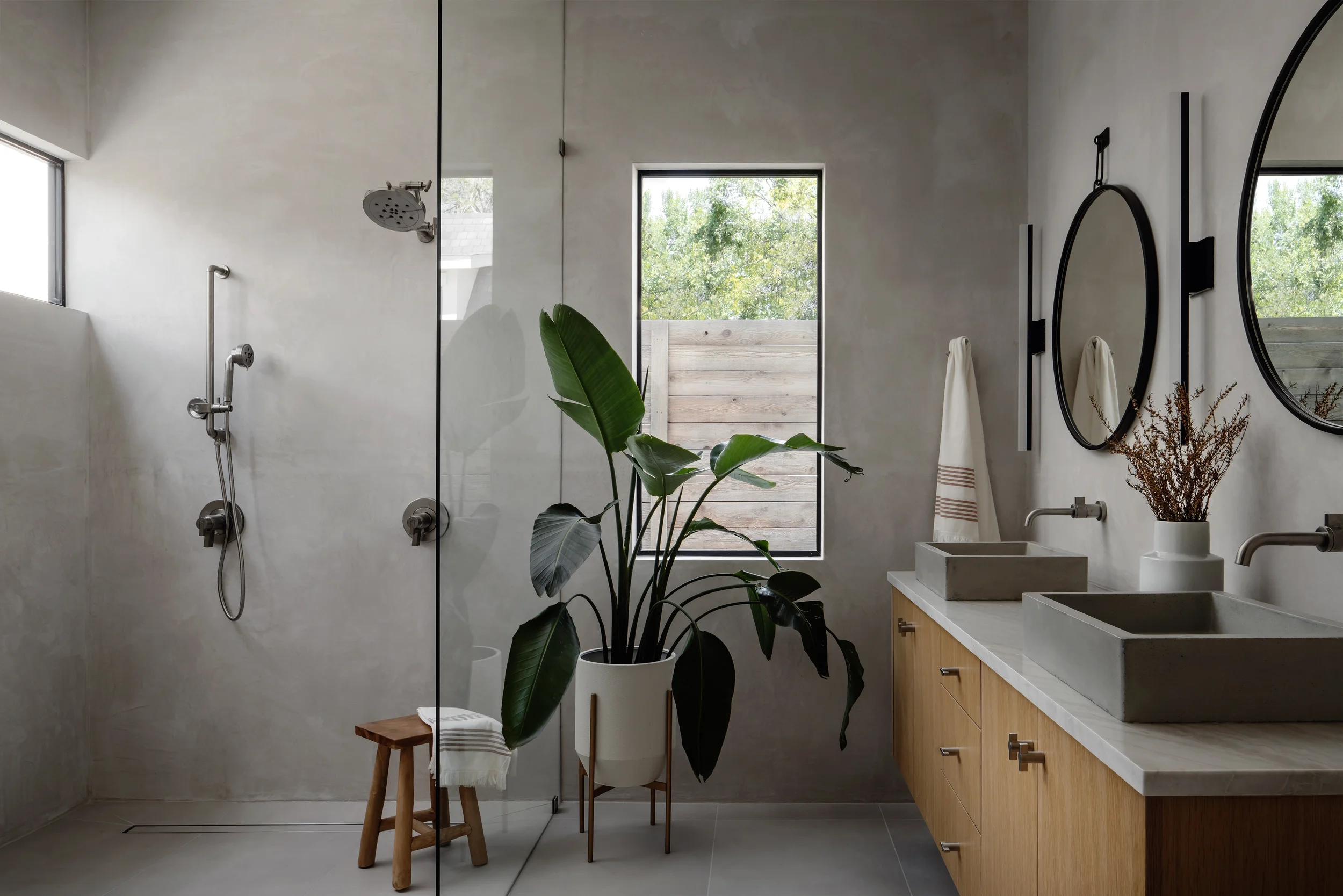 Modern bathroom with two sinks, large mirrors, a potted plant, wooden vanity, towel, and walk-in shower with glass partition and multiple showerheads.