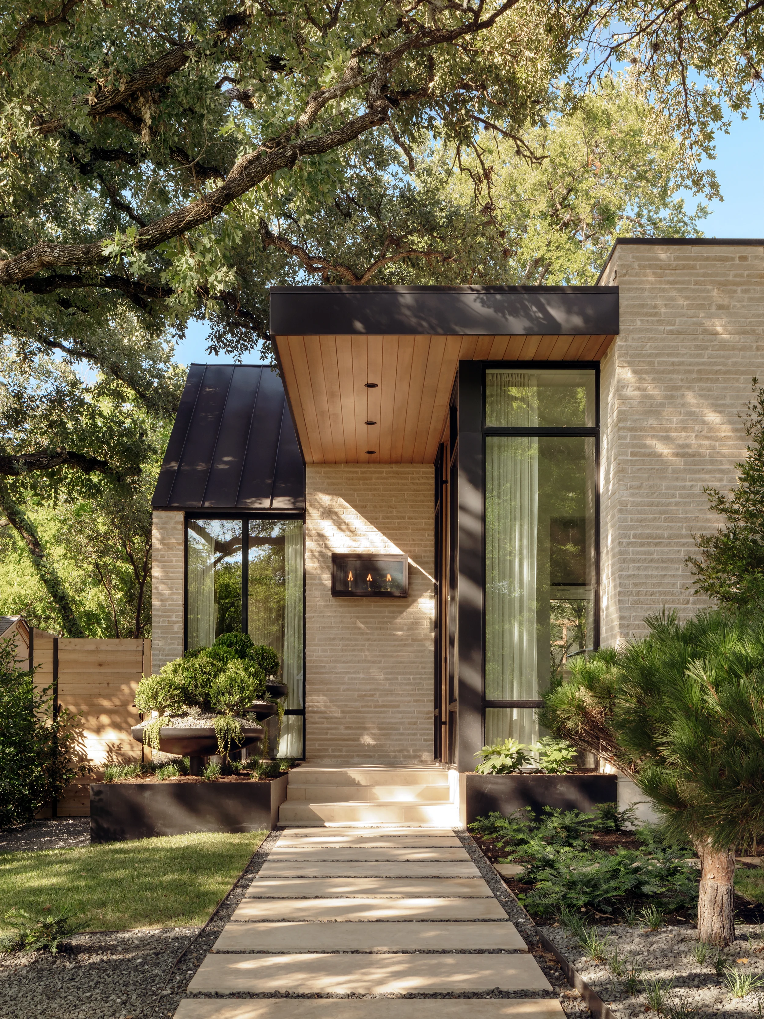 Modern house with large glass windows, metal roof accents, and a landscaped front yard with trees and shrubs.