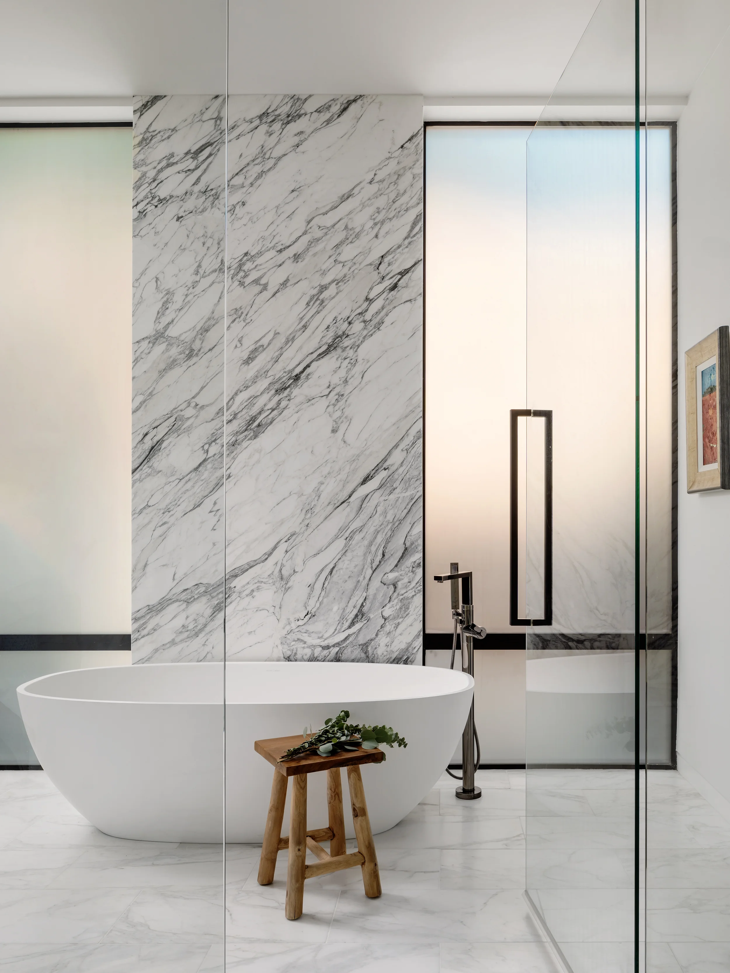 Modern bathroom with a white freestanding bathtub, marble walls, a wooden stool with greenery, and a glass shower enclosure.