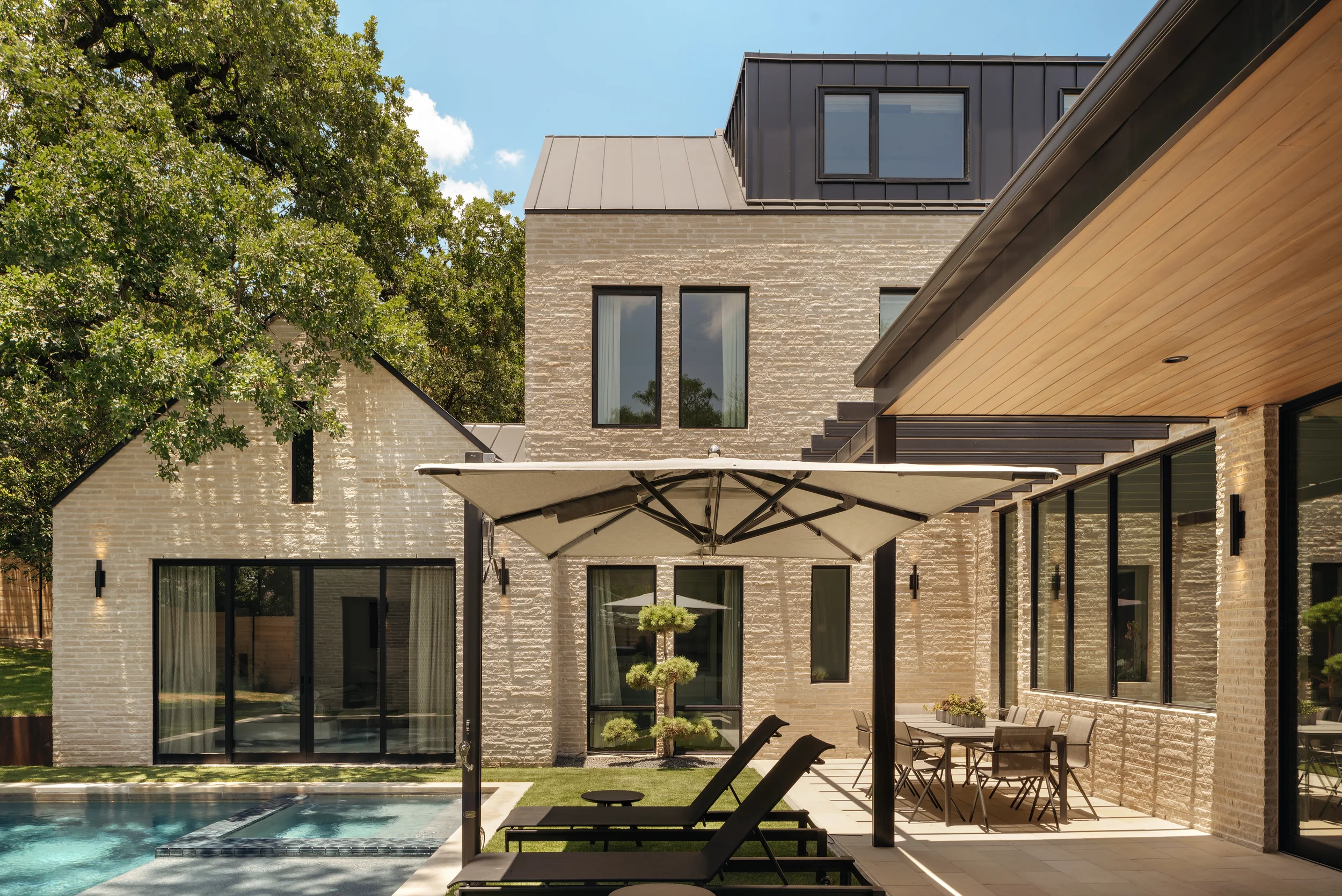 Modern house with brick exterior, outdoor patio with lounge chairs and dining table, large umbrella, swimming pool, trees, and blue sky.