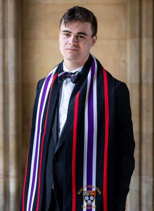 Young man in a black tuxedo with a white shirt and a bow tie, wearing a colorful scarf with red, purple, and black stripes, standing indoors against a wooden wall.