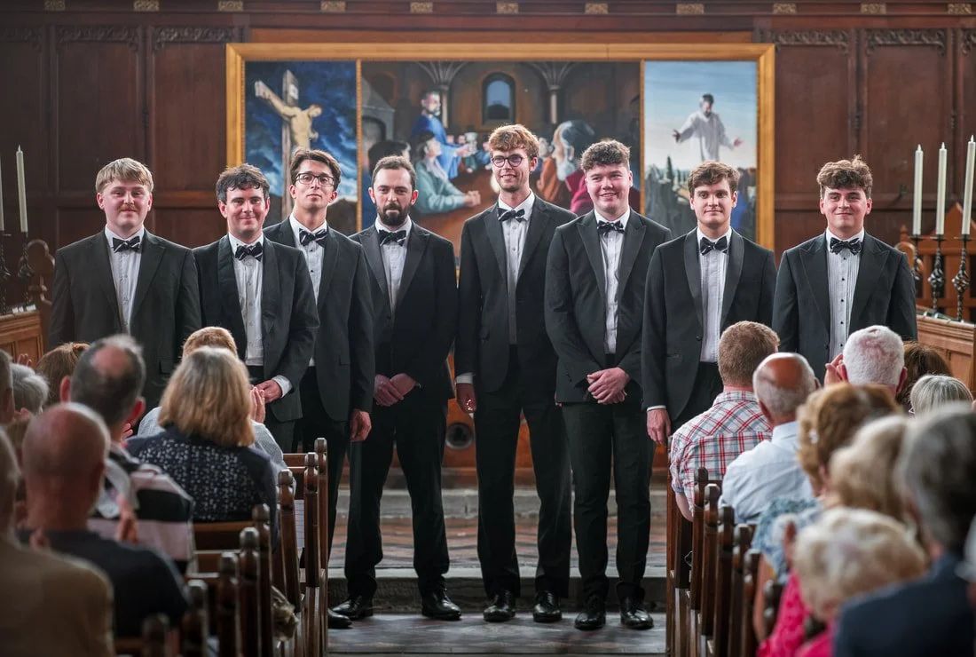A group of eight young men in tuxedos standing on a stage in a church or hall with wood paneled walls and a large religious painting behind them, addressing an audience.