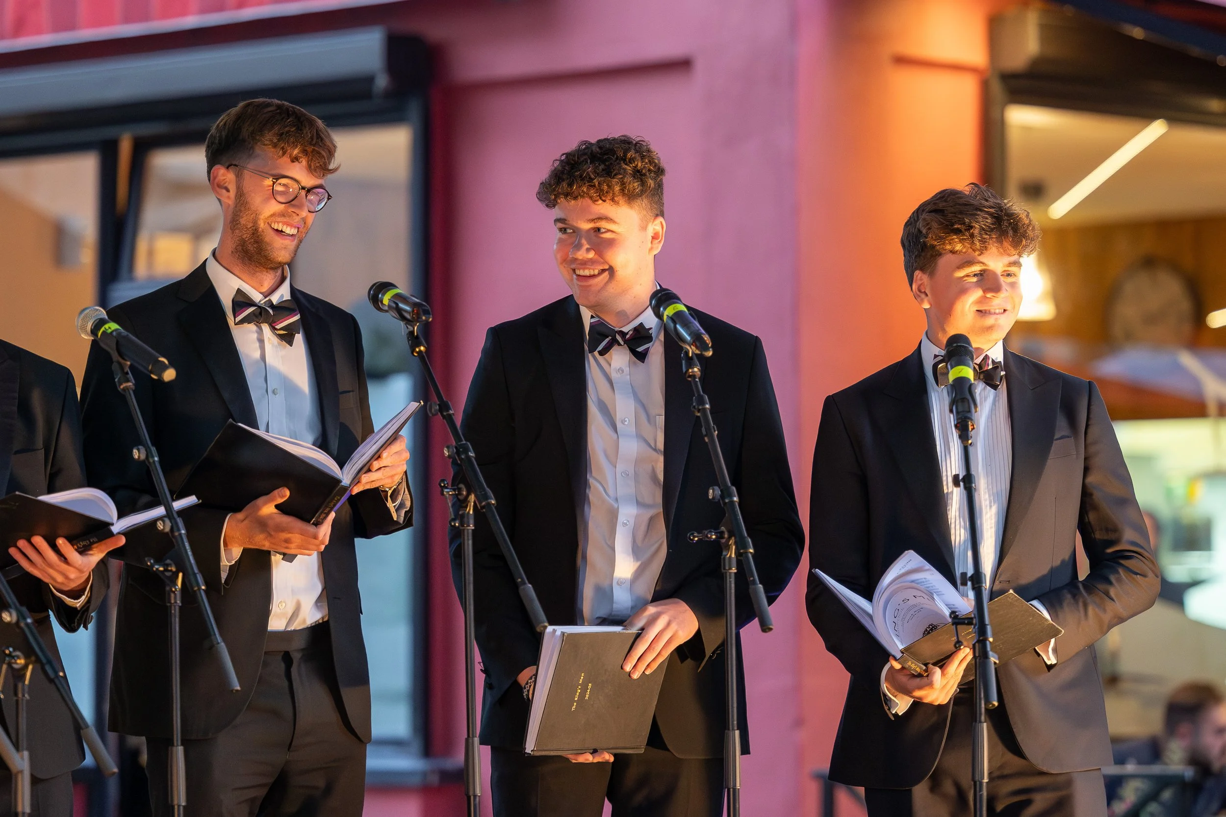 Three young men in tuxedos with bow ties, holding music books, standing on stage with microphones, smiling during a performance.
