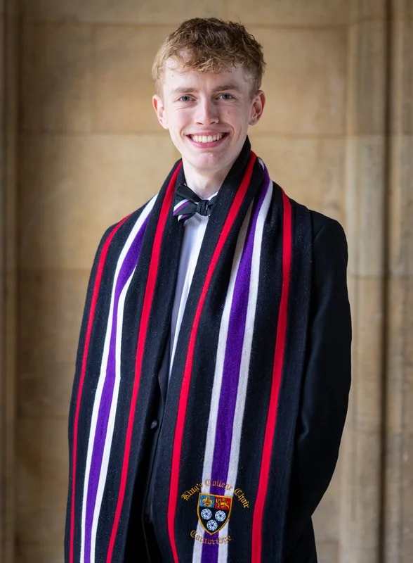 A young man dressed in a black suit, white shirt, and black bow tie, standing indoors with a stone wall background, wearing a graduation stole with black, purple, red, and white stripes and the university crest.