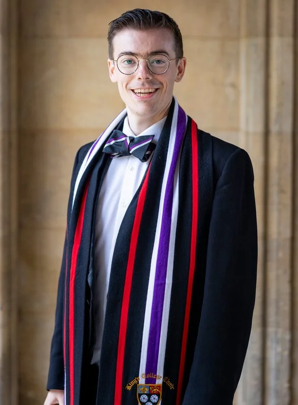 A young man in academic regalia with a black gown, a colorful scarf, and a purple and black bow tie stands in front of a stone wall, smiling at the camera.