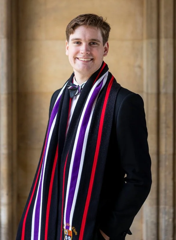 Young man in a suit wearing a black scarf with purple, white, red, and black stripes, standing in front of a stone wall, smiling at the camera.