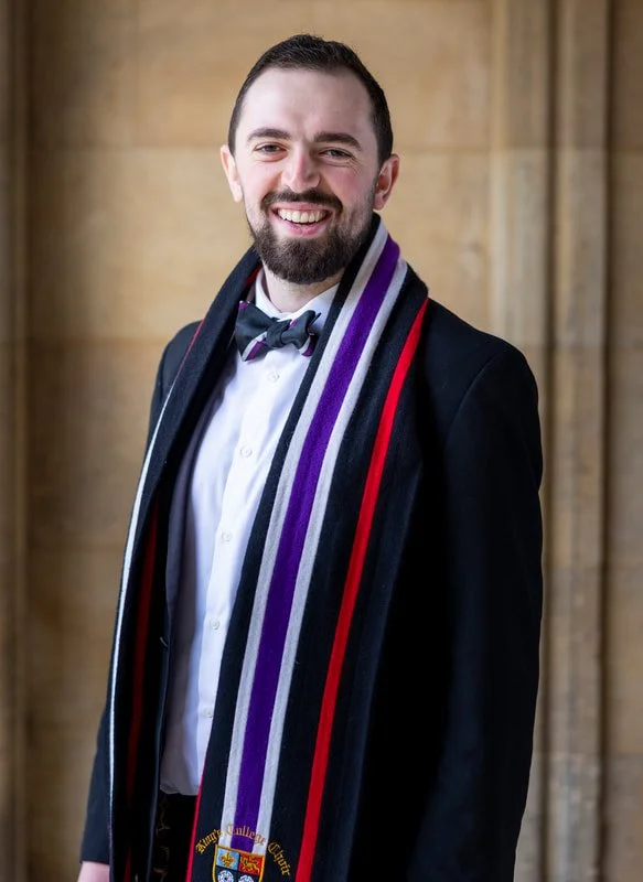 A young man dressed in formal attire, wearing a black tuxedo with a purple, black, and red academic stole and a bow tie, standing indoors with a stone wall background, smiling at the camera.